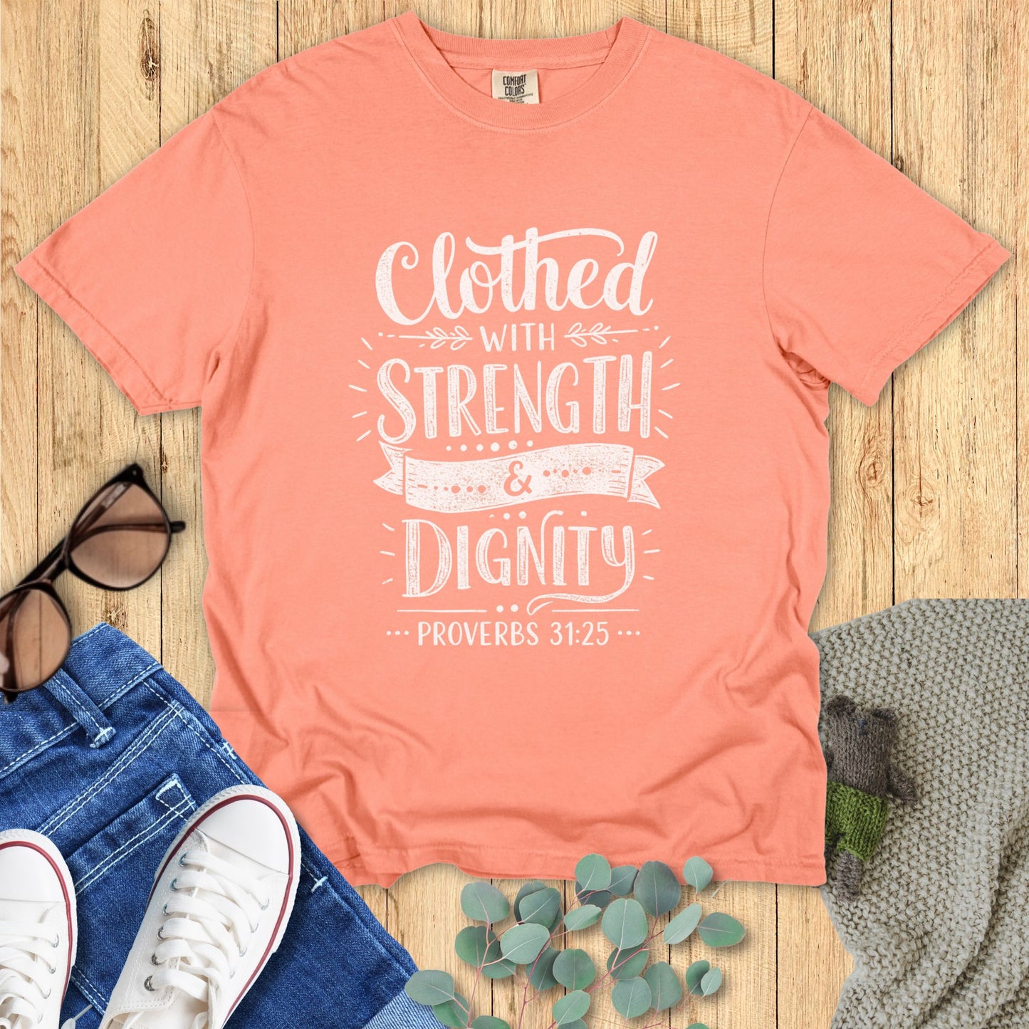 Coral t-shirt with 'Clothed with Strength & Dignity' text on a wooden surface with jeans and sneakers.