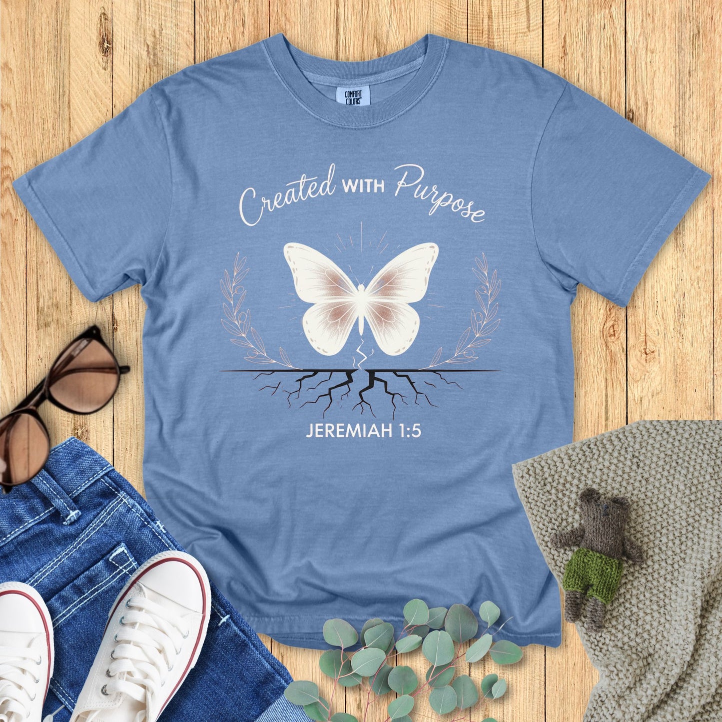 Blue t-shirt with butterfly design and 'Created with Purpose' text on a wooden surface with accessories.