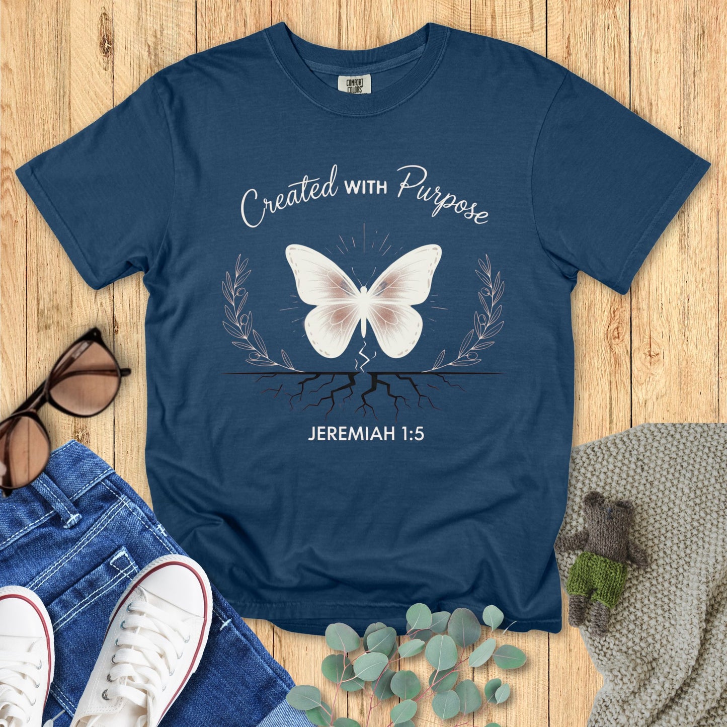 Blue t-shirt with butterfly design and 'Created with Purpose' text on a wooden surface with accessories.