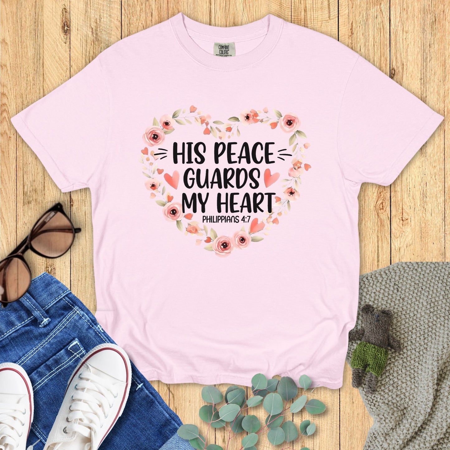 Pink t-shirt with floral design and text on a wooden surface with accessories.