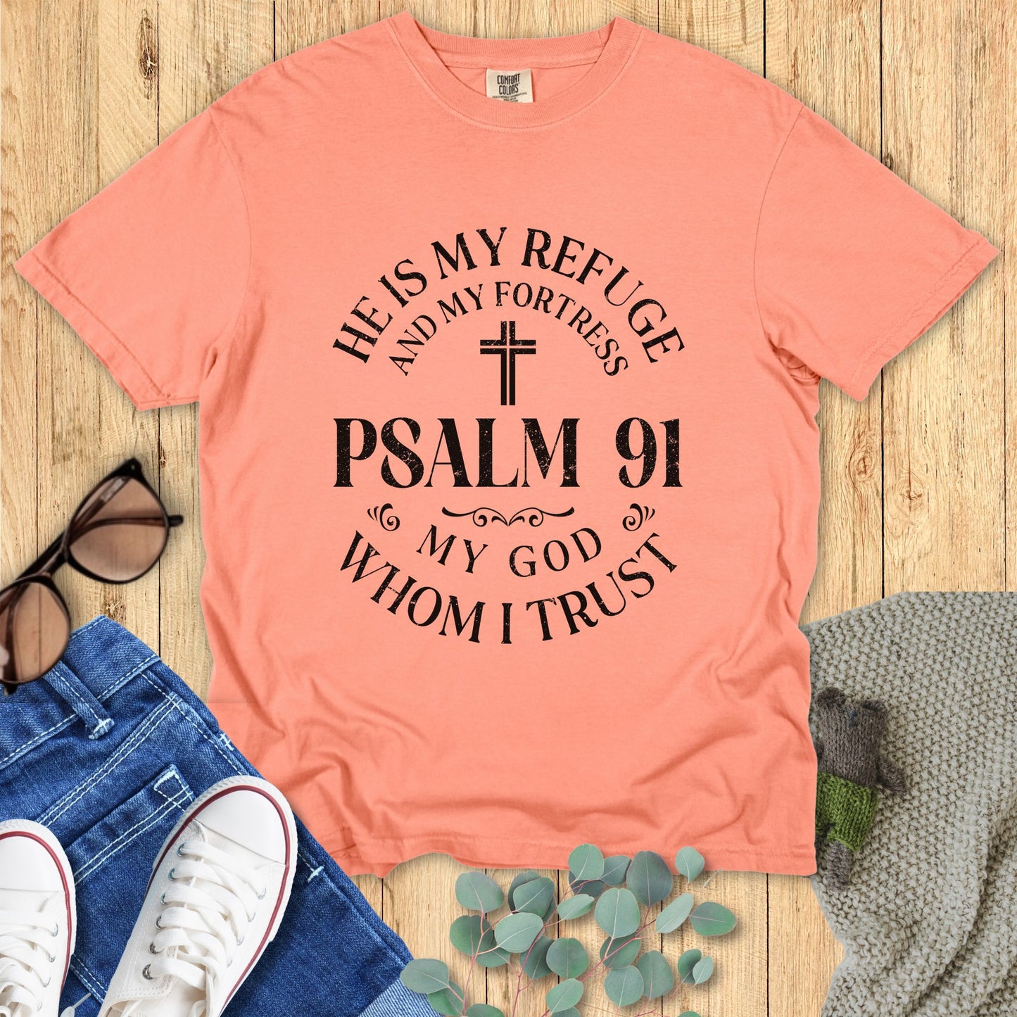 Peach t-shirt with 'He is my refuge and my fortress, Psalm 91' text on a wooden surface with jeans and sneakers.