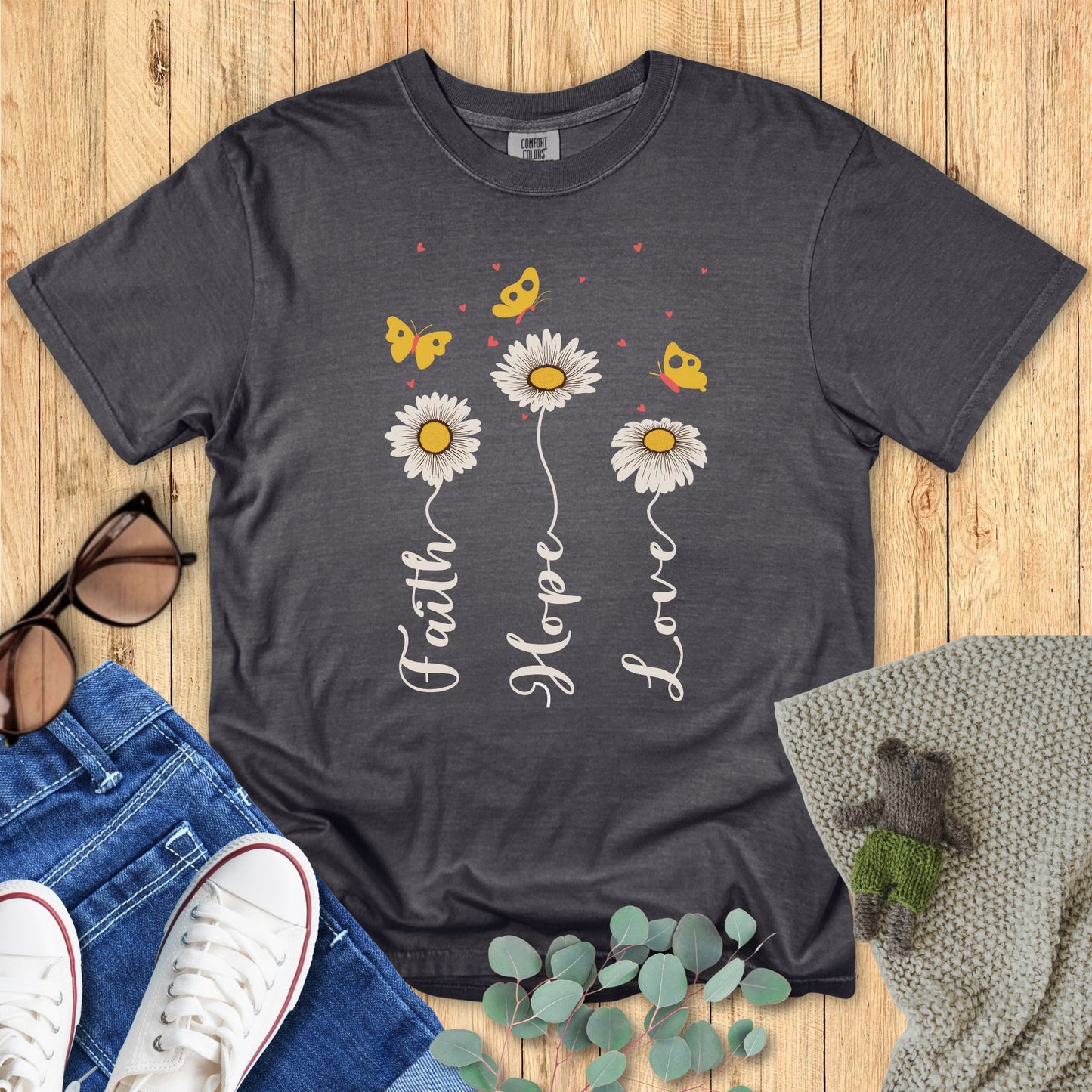 Gray t-shirt with floral design and text on a wooden surface with accessories.