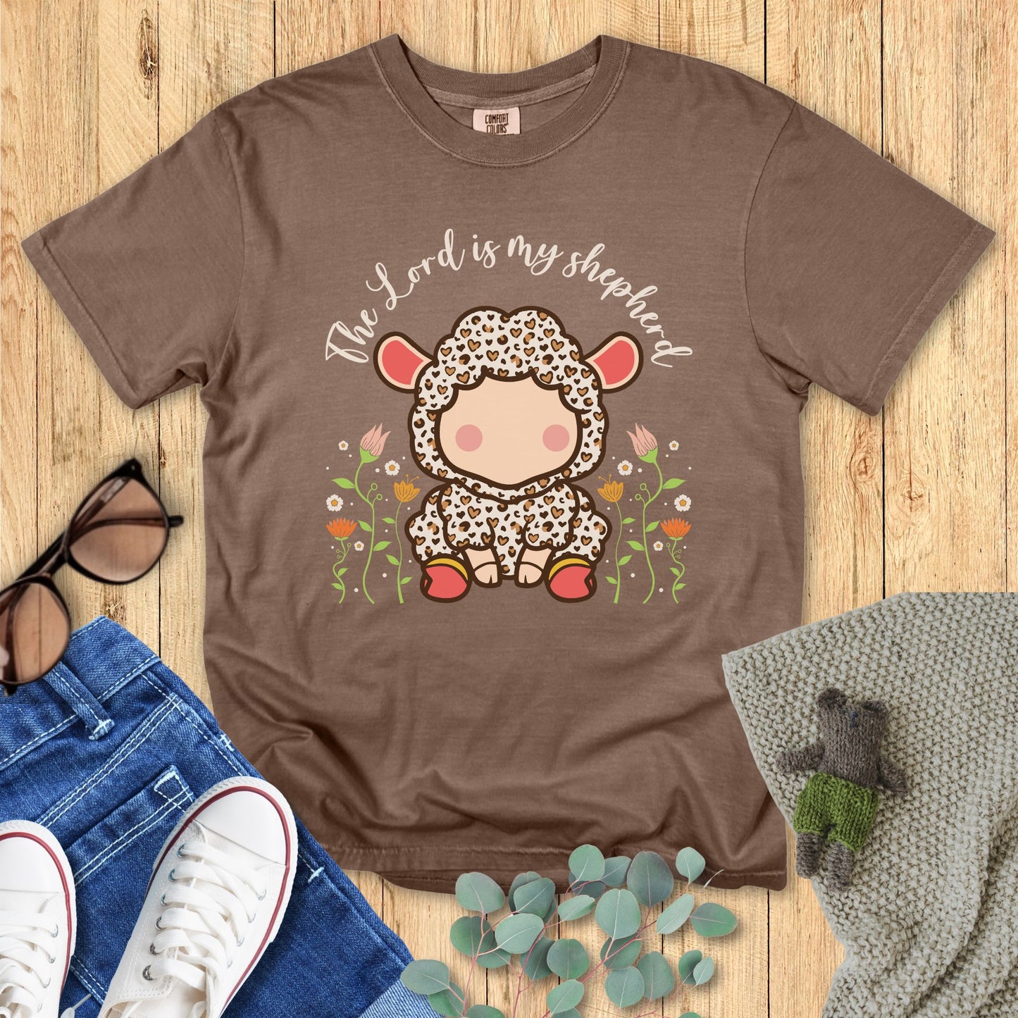 Brown t-shirt with a sheep graphic and 'The Lord is my shepherd' text on a wooden surface with accessories.