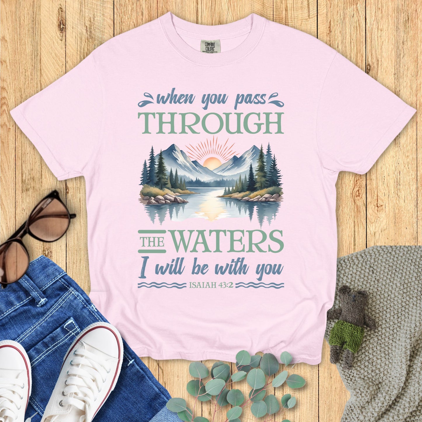 Pink t-shirt with nature graphic and text on a wooden surface with accessories.