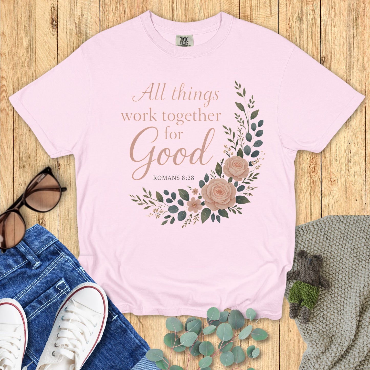 Pink t-shirt with floral design and text on a wooden surface with jeans and sneakers.