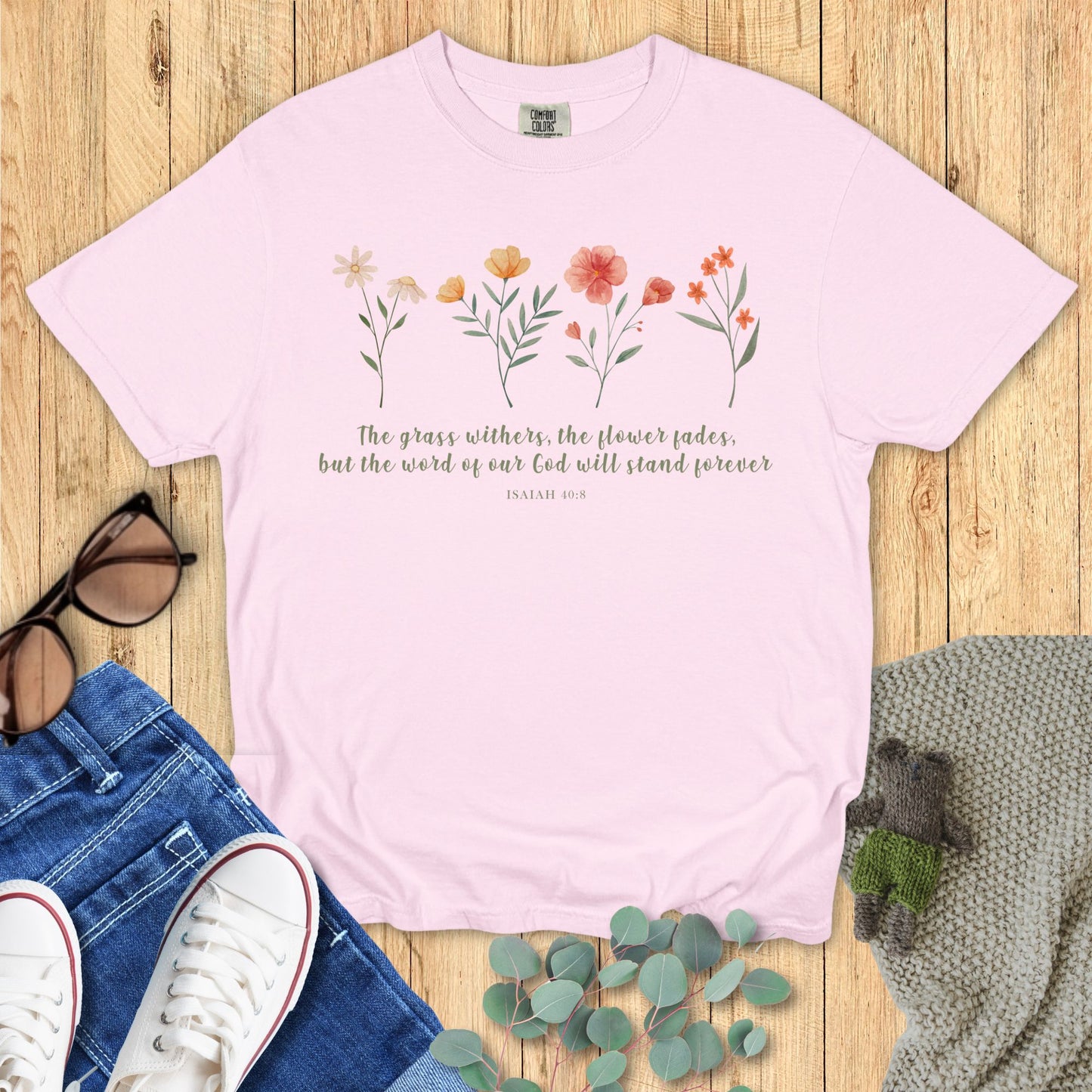 Pink t-shirt with floral design and text on a wooden surface with jeans and sneakers.