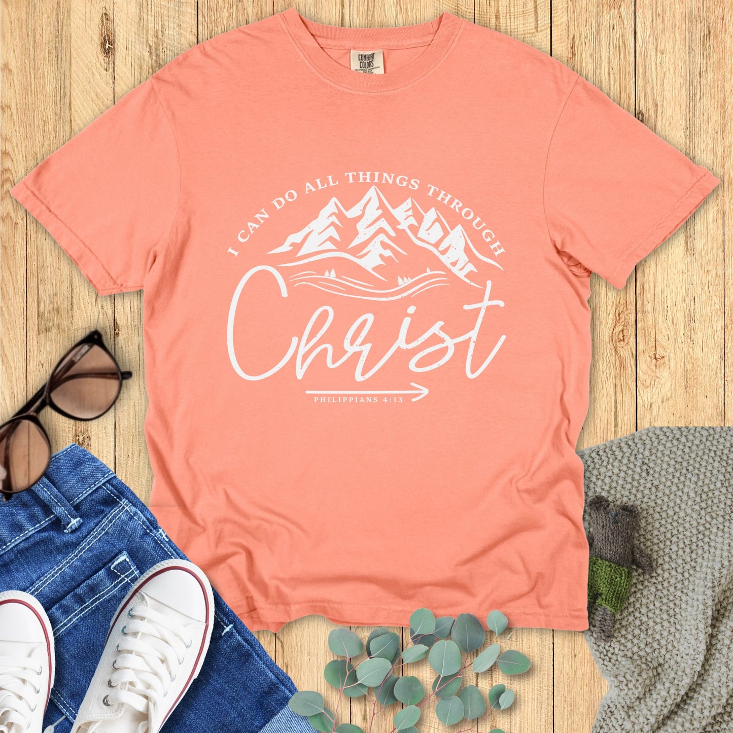 Coral t-shirt with motivational quote on a wooden surface with accessories.