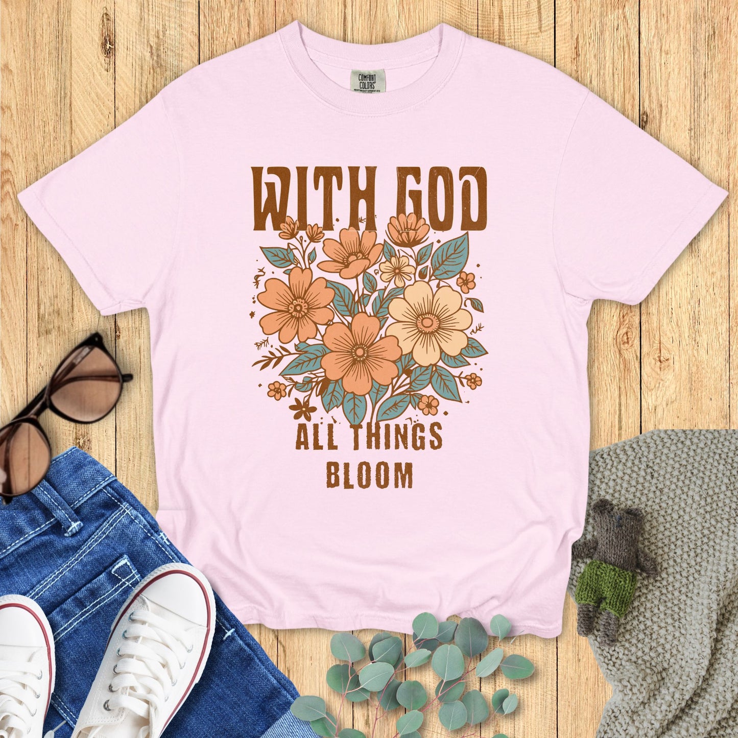 Pink t-shirt with floral design and text on a wooden surface with accessories.