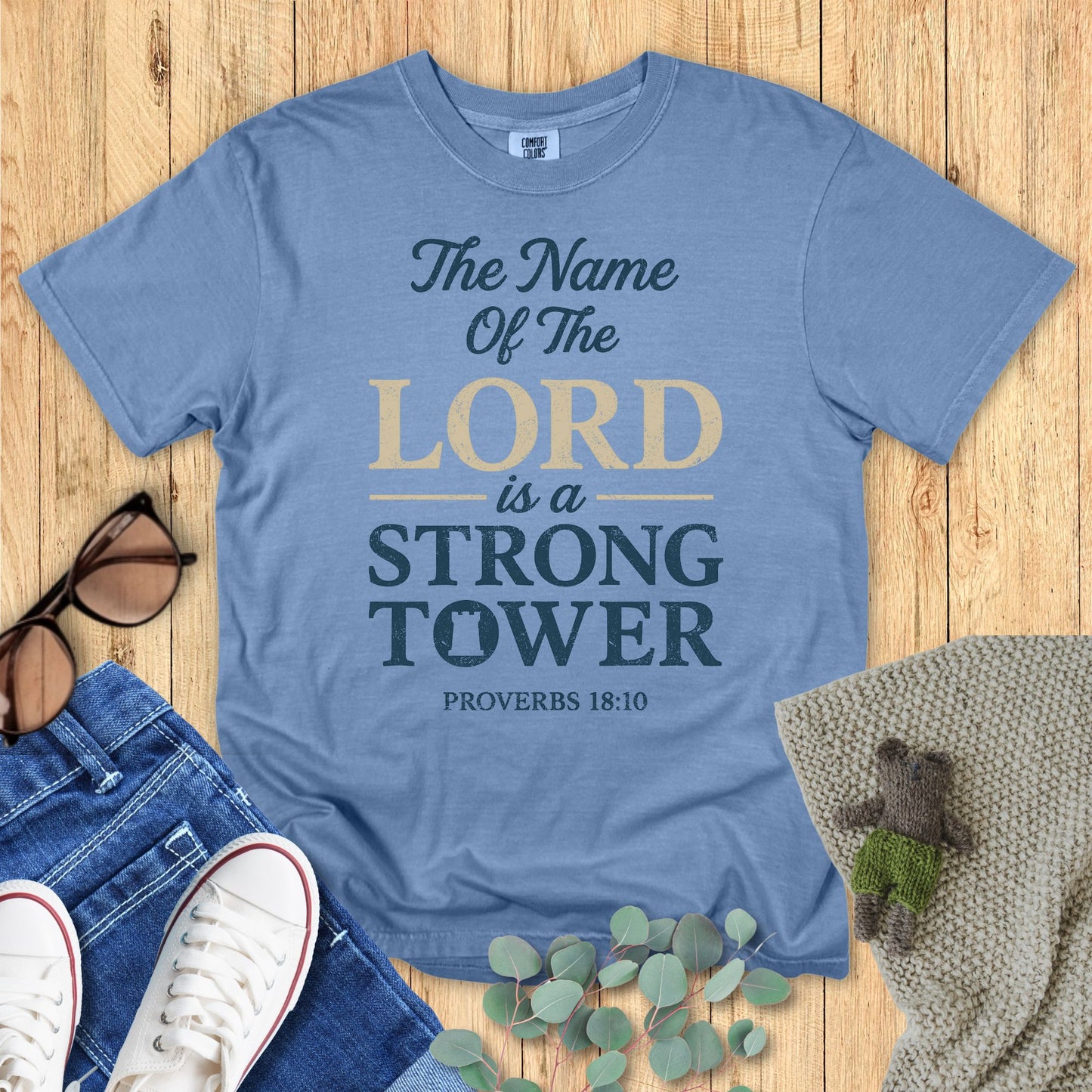 Blue t-shirt with 'The Name of the Lord is a Strong Tower' text on a wooden surface with accessories.