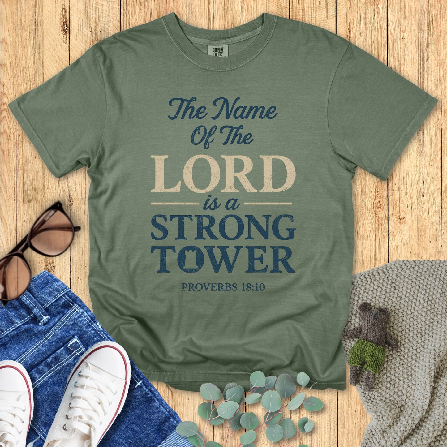 Green t-shirt with 'The Name of the Lord is a Strong Tower' text on a wooden surface with accessories.