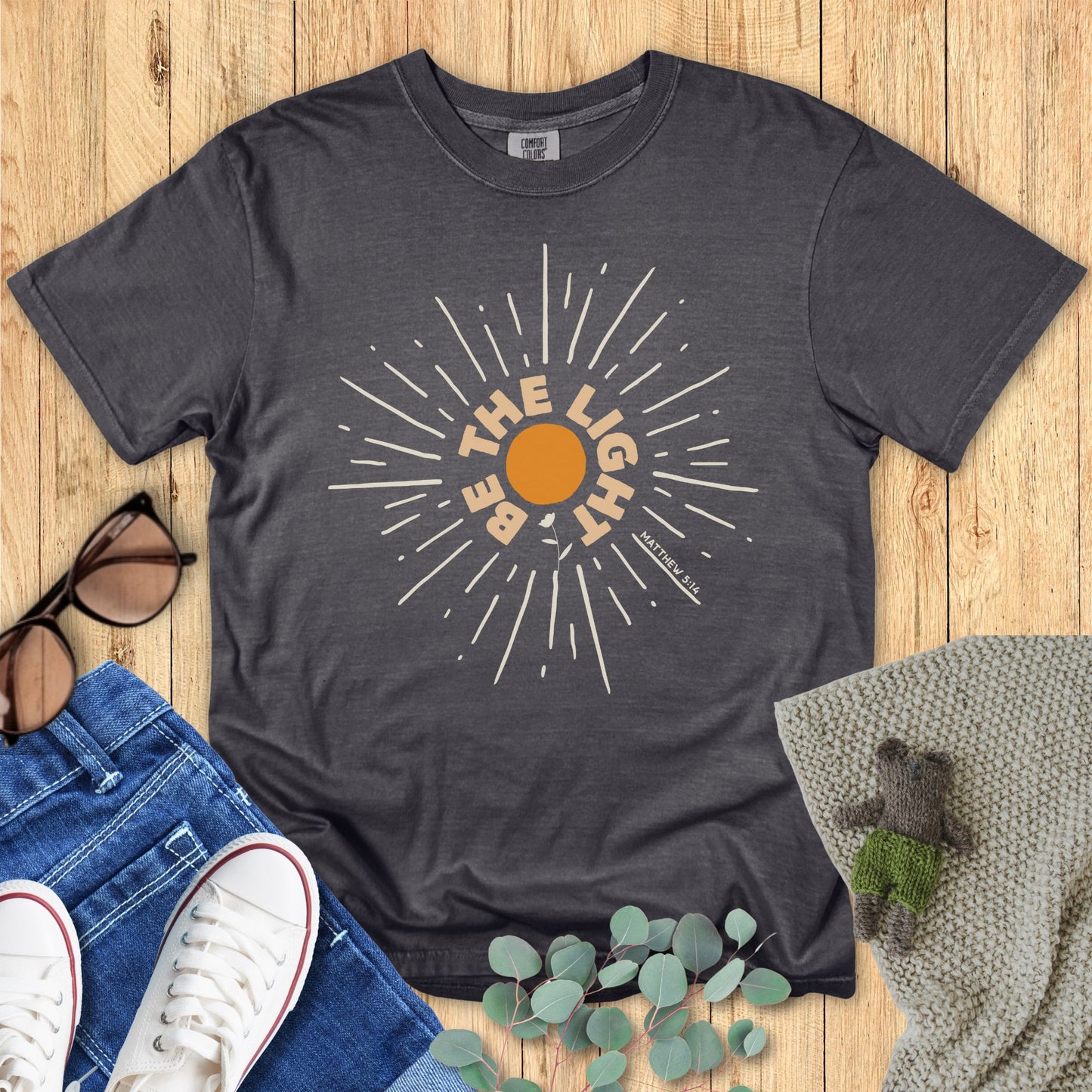 Gray t-shirt with sun graphic and text on a wooden surface with accessories.