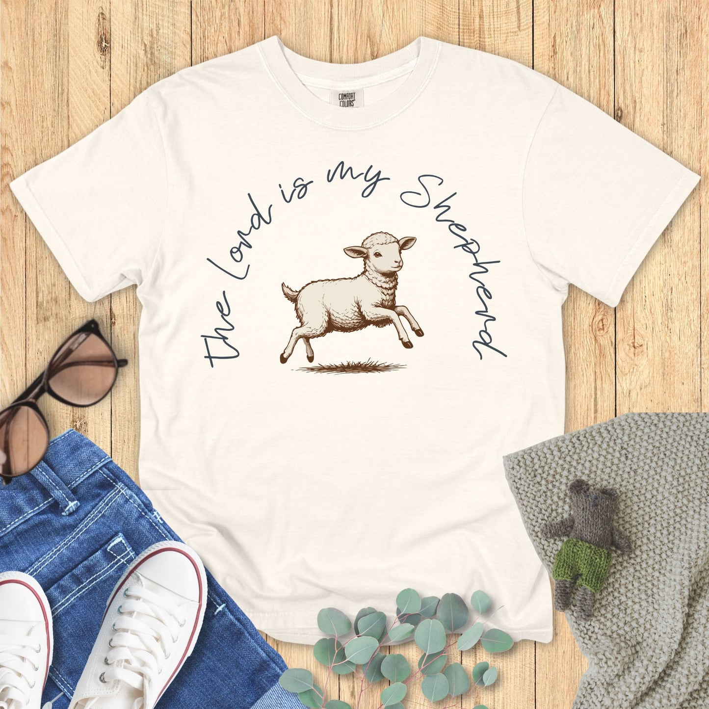 White t-shirt with a sheep graphic and text on a wooden surface with accessories.