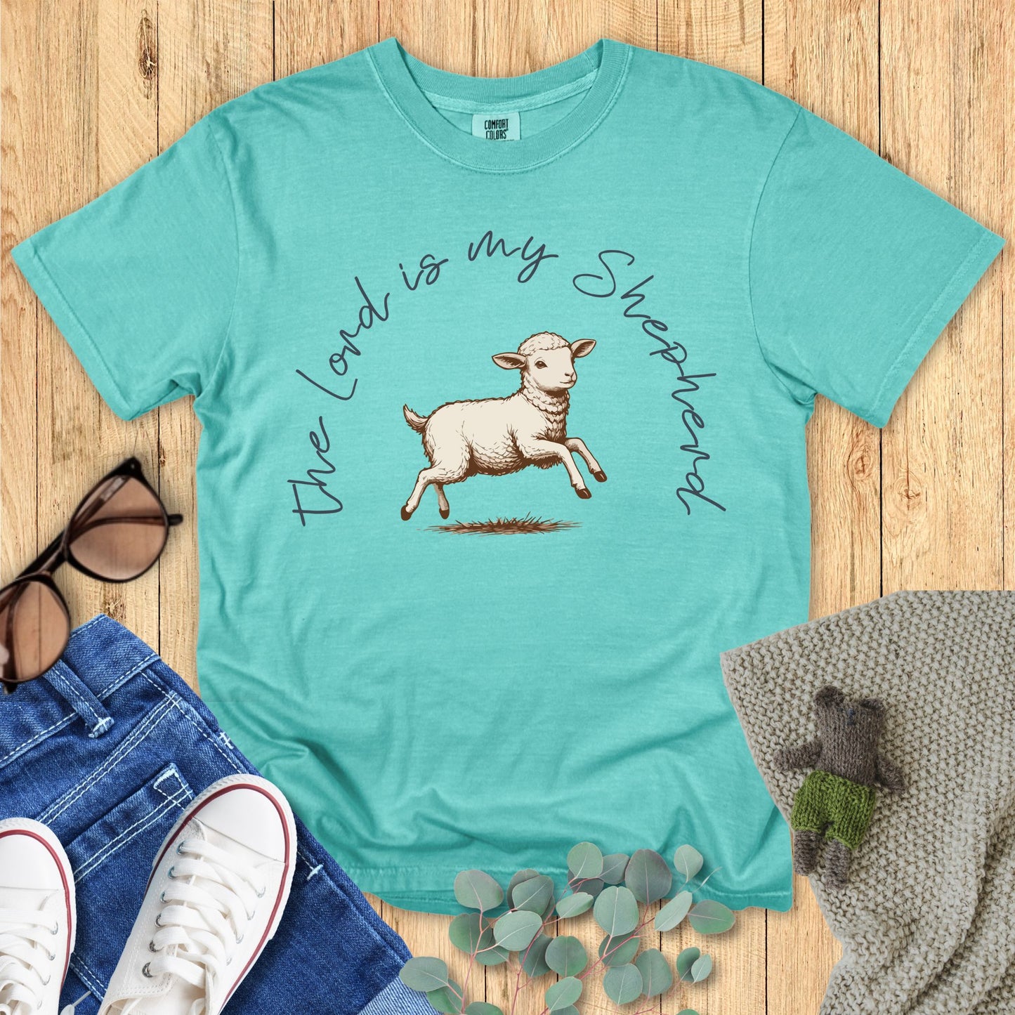 Teal t-shirt with a sheep graphic and text on a wooden surface with accessories.