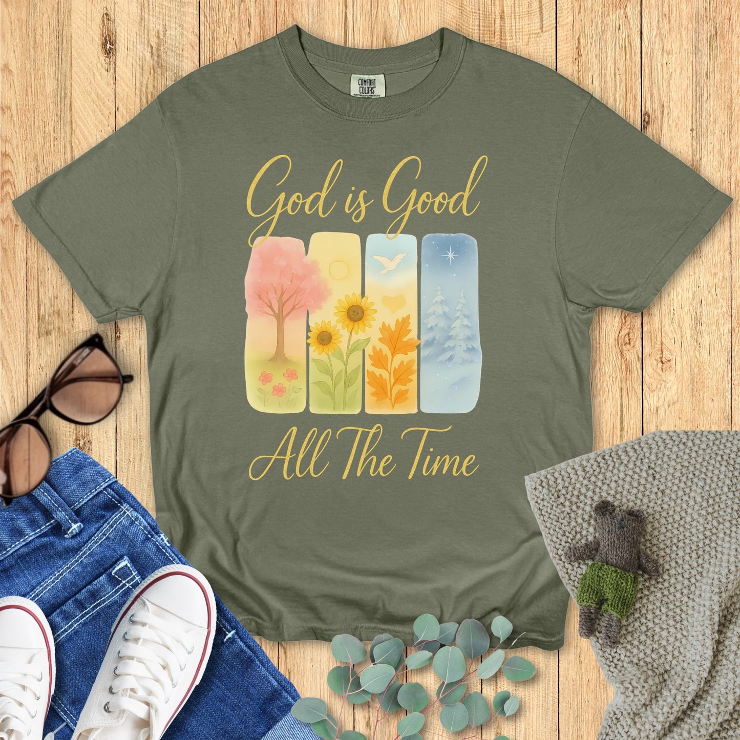 Green t-shirt with 'God is Good All The Time' design on a wooden surface with accessories.