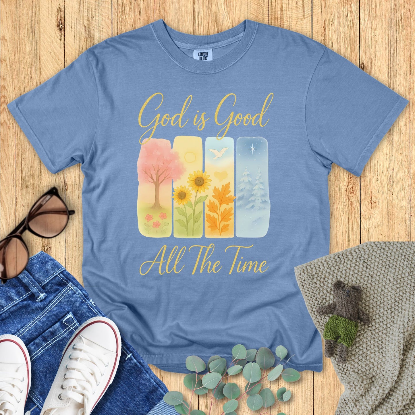 Blue t-shirt with 'God is Good All The Time' text and nature-themed design on a wooden surface with accessories.