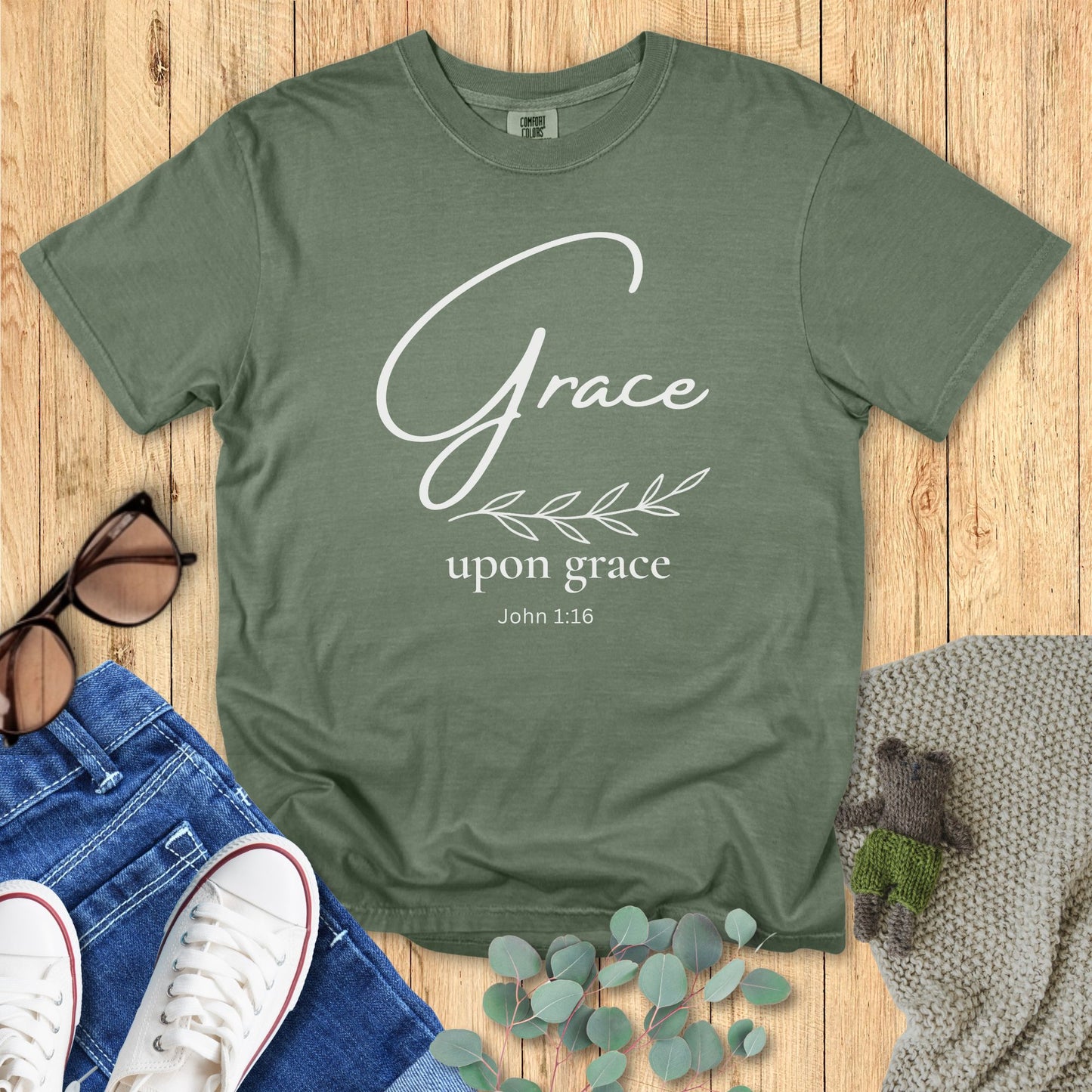 Green t-shirt with 'Grace upon grace John 1:16' text on a wooden surface with accessories.