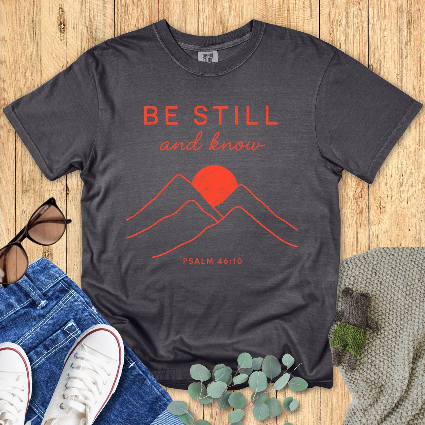 Gray t-shirt with red mountain design and text on a wooden surface with jeans and sunglasses.