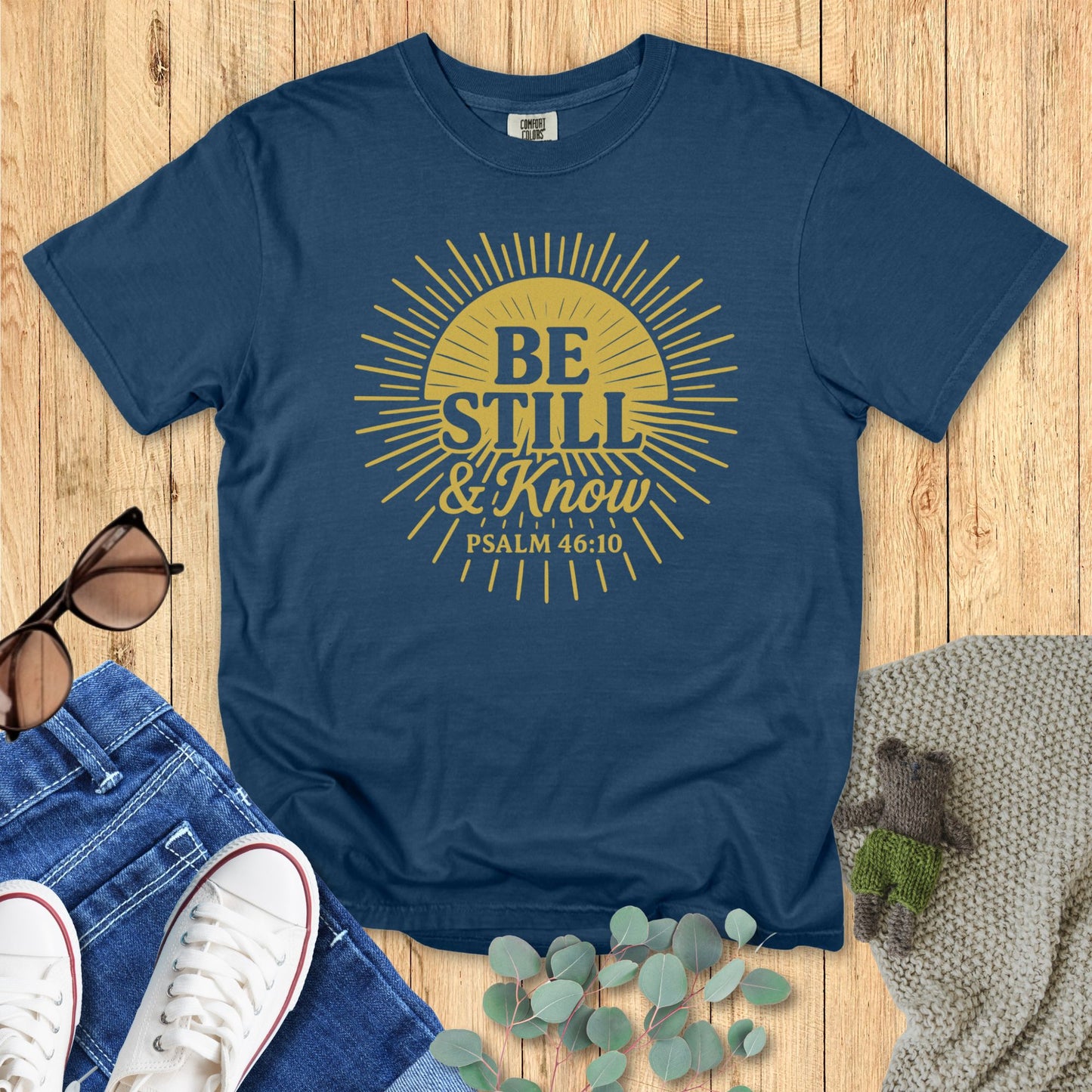 Blue t-shirt with yellow text 'Be Still & Know Psalm 46:10' on a wooden surface with jeans and sneakers.