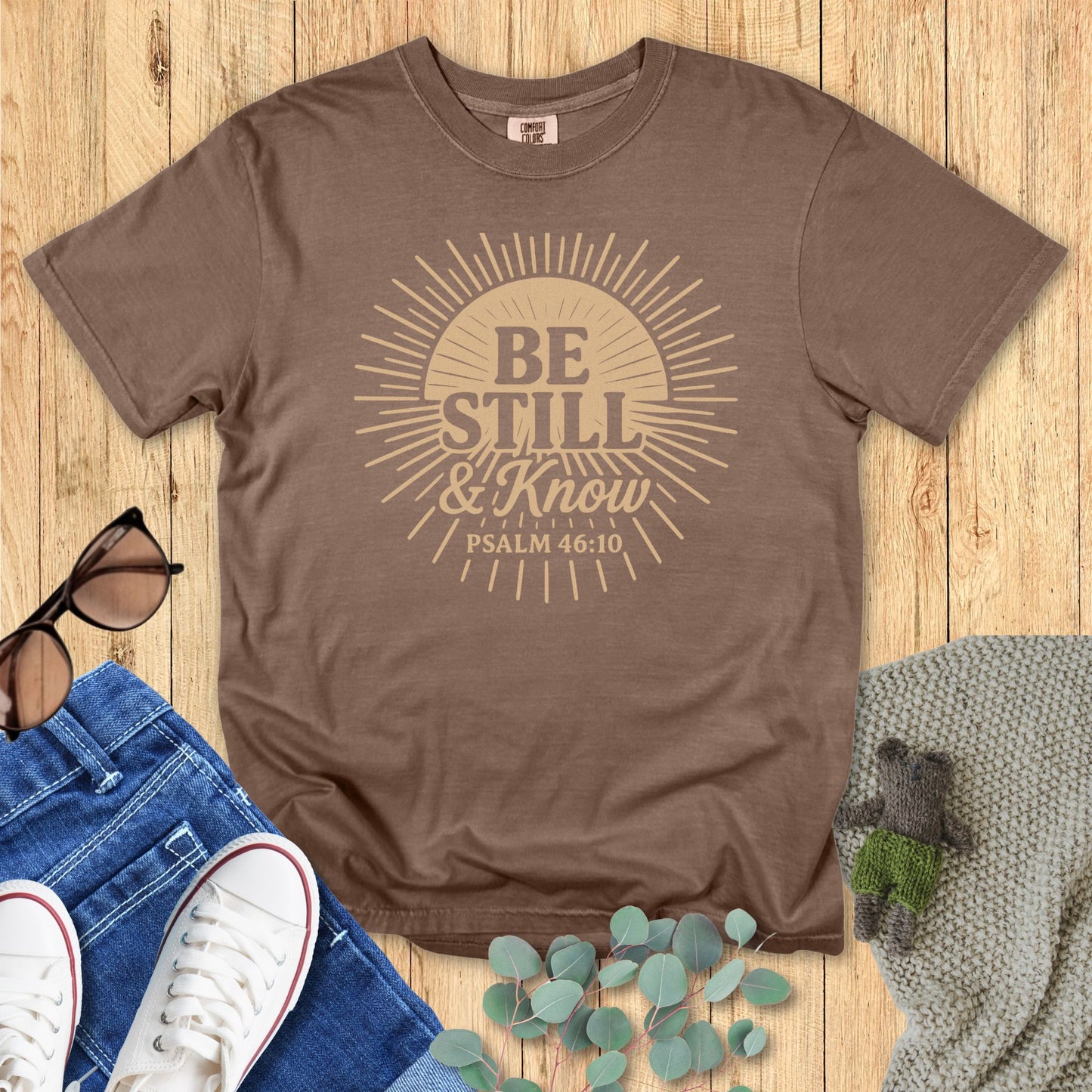 Brown t-shirt with 'Be Still & Know' design on a wooden surface with jeans, sneakers, and leaves.
