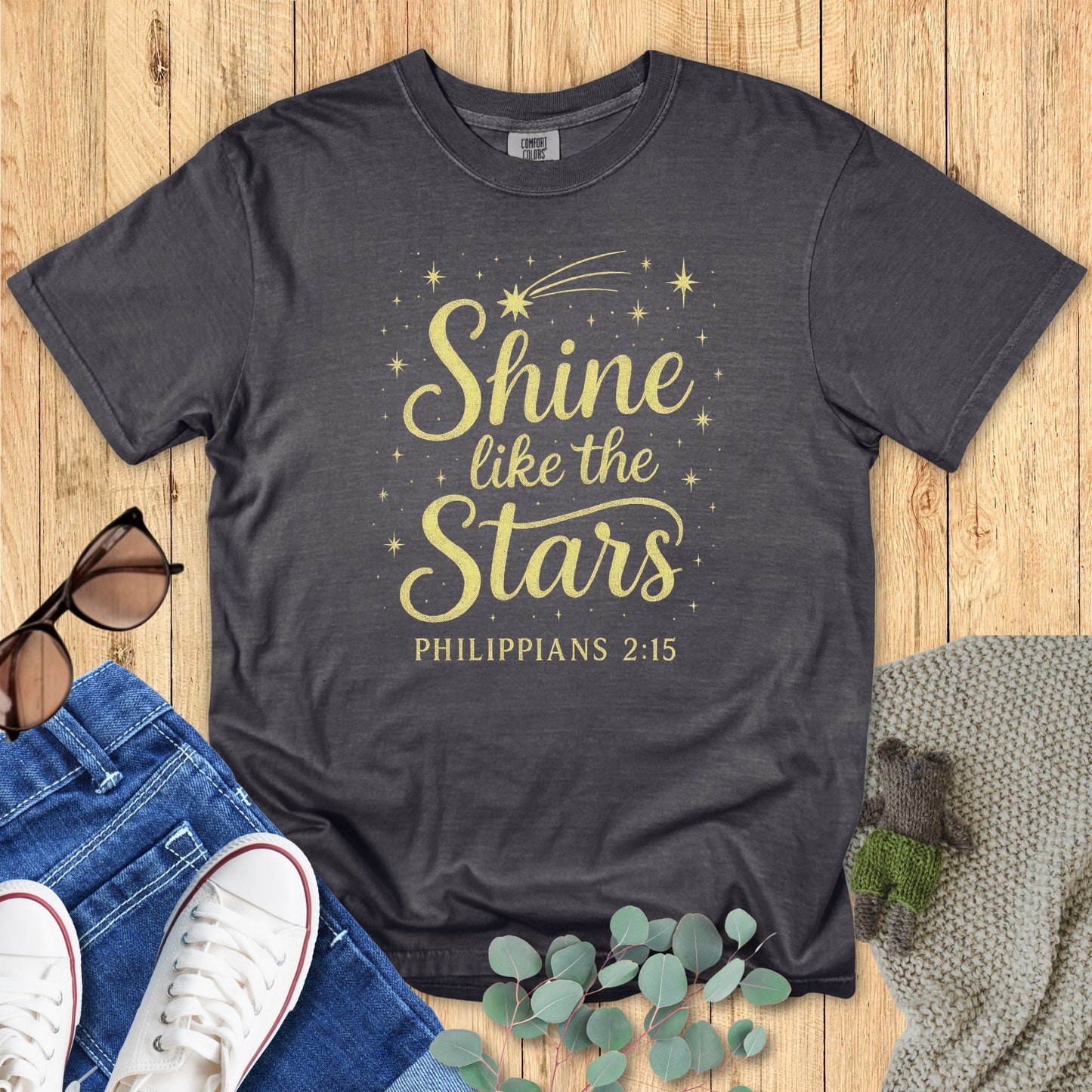 Gray t-shirt with 'Shine like the stars' text on a wooden surface with accessories.