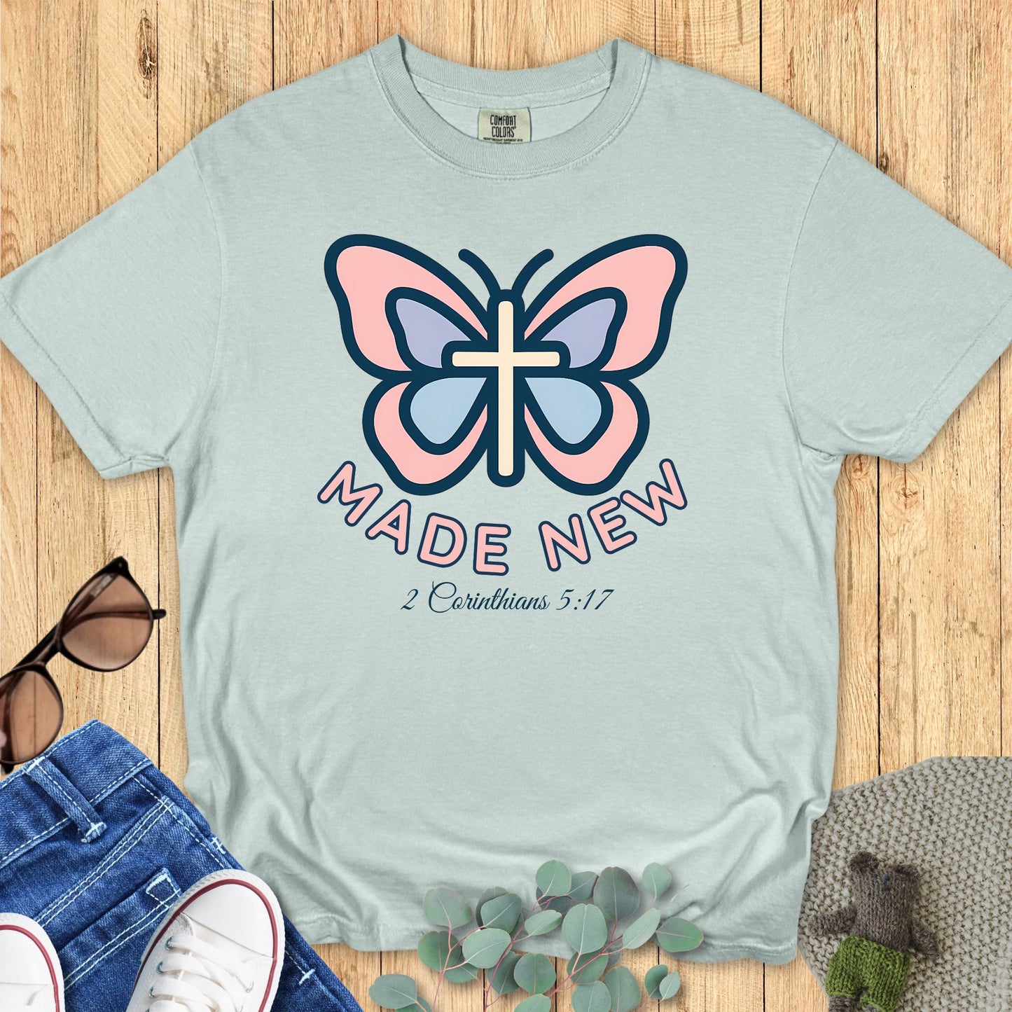 Comfort Colors 1717 Bay t-shirt with a vibrant pastel butterfly and cross graphic, "Made New" text, and 2 Corinthians 5:17 Bible verse below.