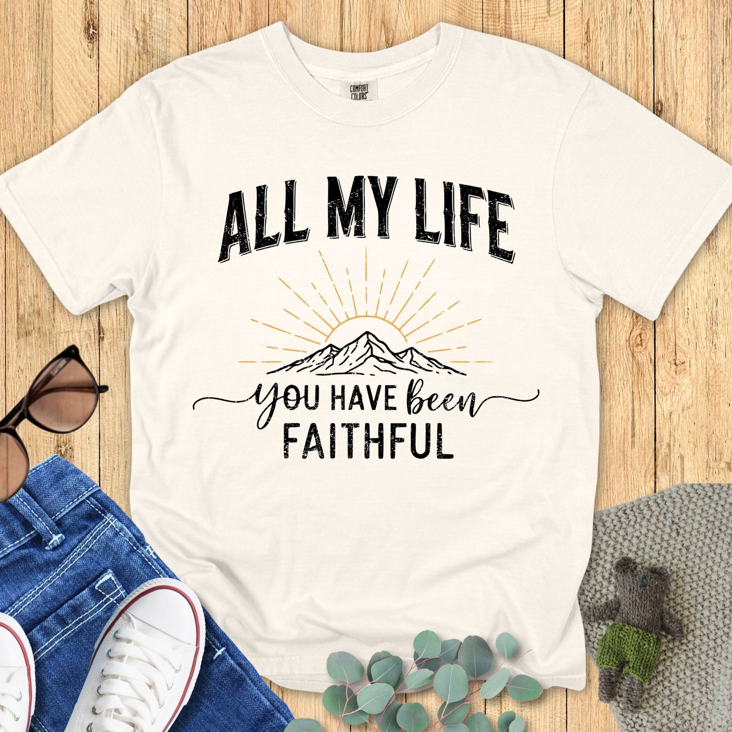 A white ivory Comfort Colors T-shirt laid flat on a wooden background. The shirt design features the phrase “ALL MY LIFE” in bold distressed black uppercase letters above a hand-drawn mountain illustration with golden sun rays. Below the mountain, the text reads “you have been faithful” in a mix of script and bold block font. Styled with sunglasses, denim jeans, white sneakers, and eucalyptus leaves.