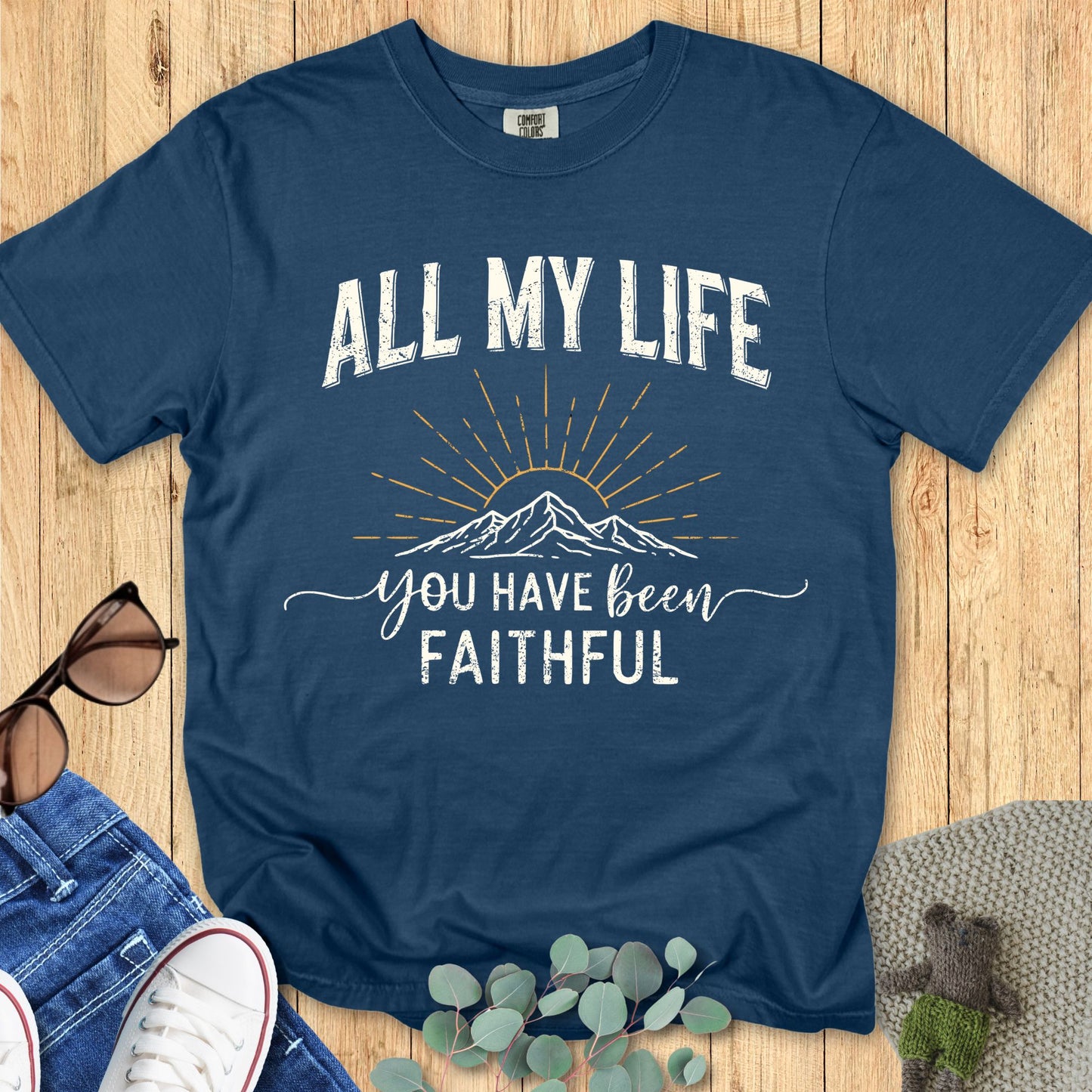 A deep midnight blue Comfort Colors T-shirt on a wooden background. The design shows the phrase “ALL MY LIFE” in distressed white uppercase letters above a sketched mountain with golden sun rays. The words “you have been faithful” are printed below in a mix of cursive and block font. Accessories include sunglasses, denim jeans, white sneakers, and eucalyptus leaves for a casual styled look.