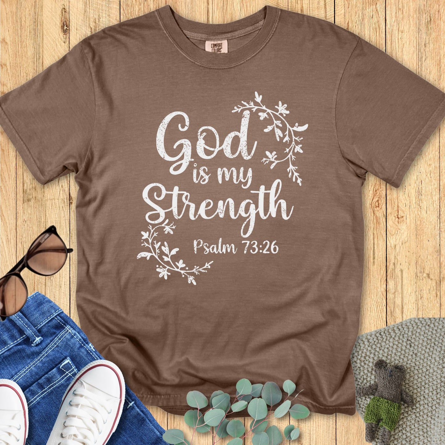 Brown Comfort Colors T-shirt with white script text that reads “God is my Strength – Psalm 73:26,” accented by delicate floral vine illustrations around the wording. Styled flat lay with sunglasses, denim jeans, white sneakers, and greenery on a wooden background.