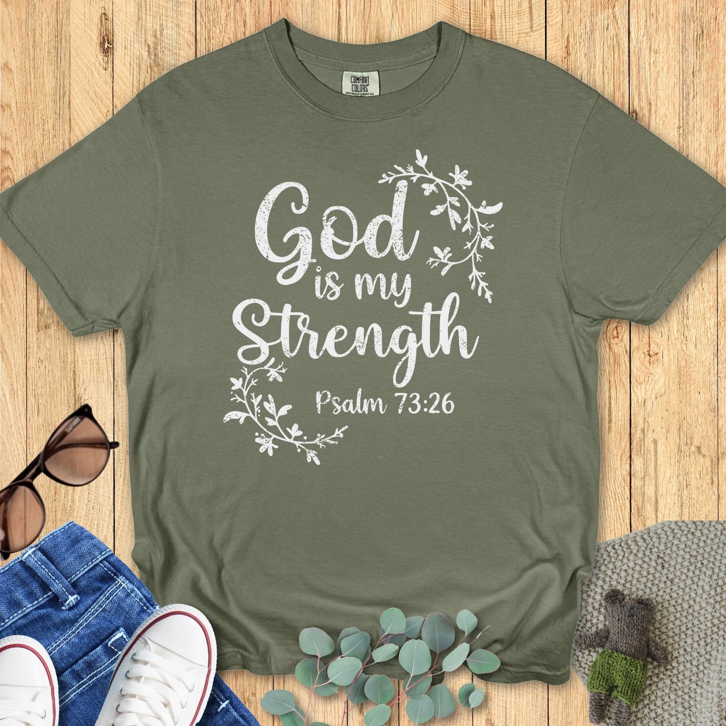 Sage green Comfort Colors T-shirt featuring white lettering that says “God is my Strength – Psalm 73:26,” framed with light floral vine accents. Flat lay display with sunglasses, denim jeans, white sneakers, and eucalyptus leaves on a wooden background.