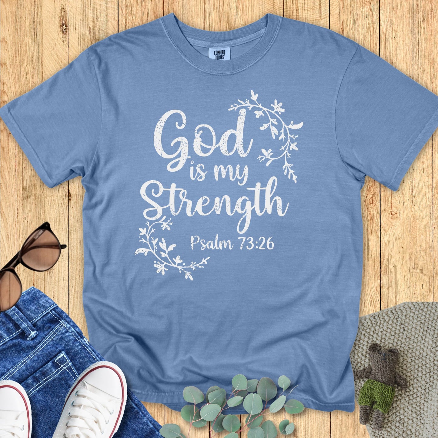 Light blue Comfort Colors T-shirt with distressed white text that reads “God is my Strength – Psalm 73:26,” decorated with floral vine graphics. Flat lay setup with sunglasses, denim jeans, white sneakers, and greenery on a wooden surface.