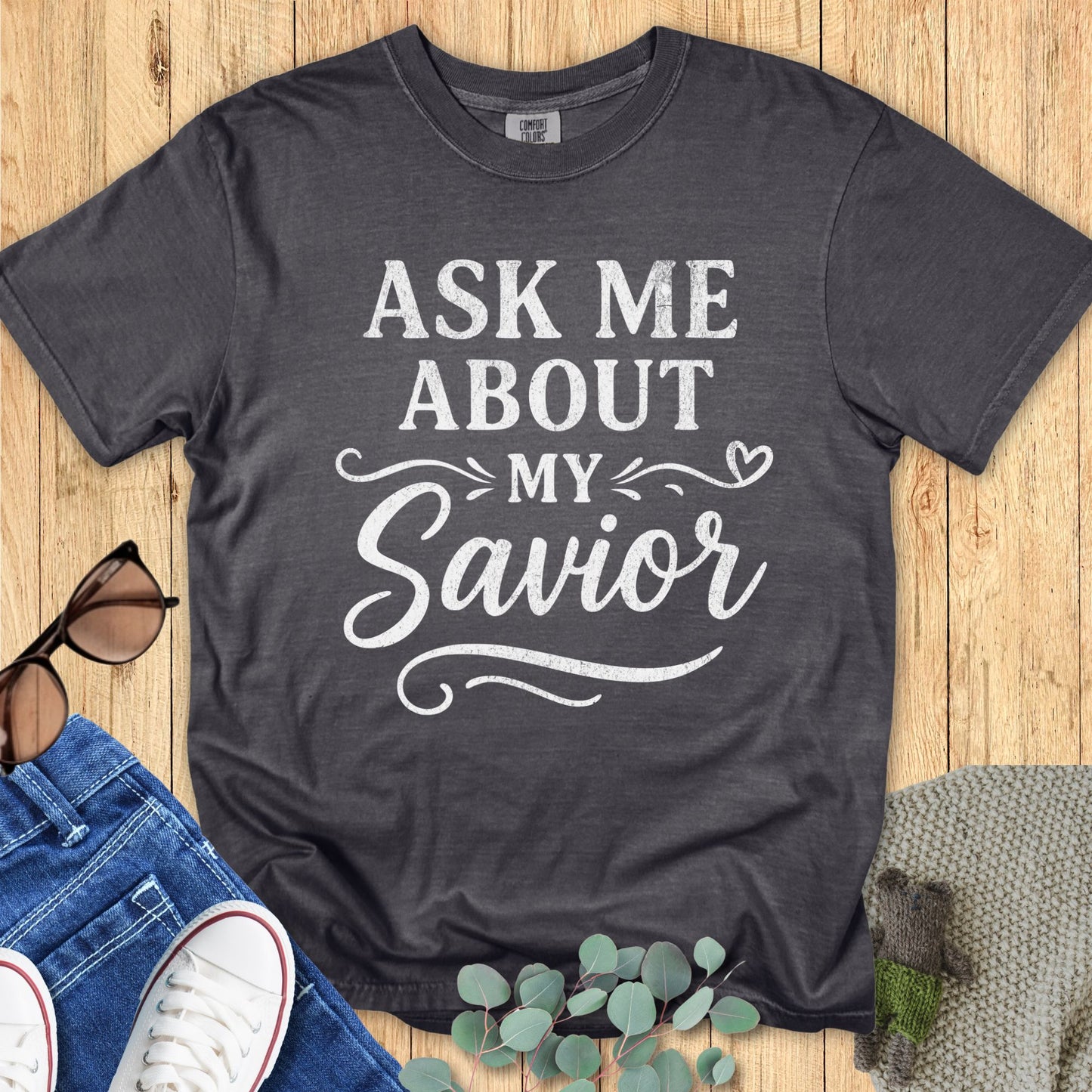 Pepper gray Comfort Colors 1717 T-shirt featuring white distressed text “Ask Me About My Savior” in bold and script lettering, accented by swirls and a small heart. A meaningful shirt that lets you wear your faith with confidence. Displayed flat on wood with jeans, sneakers, sunglasses, and eucalyptus leaves