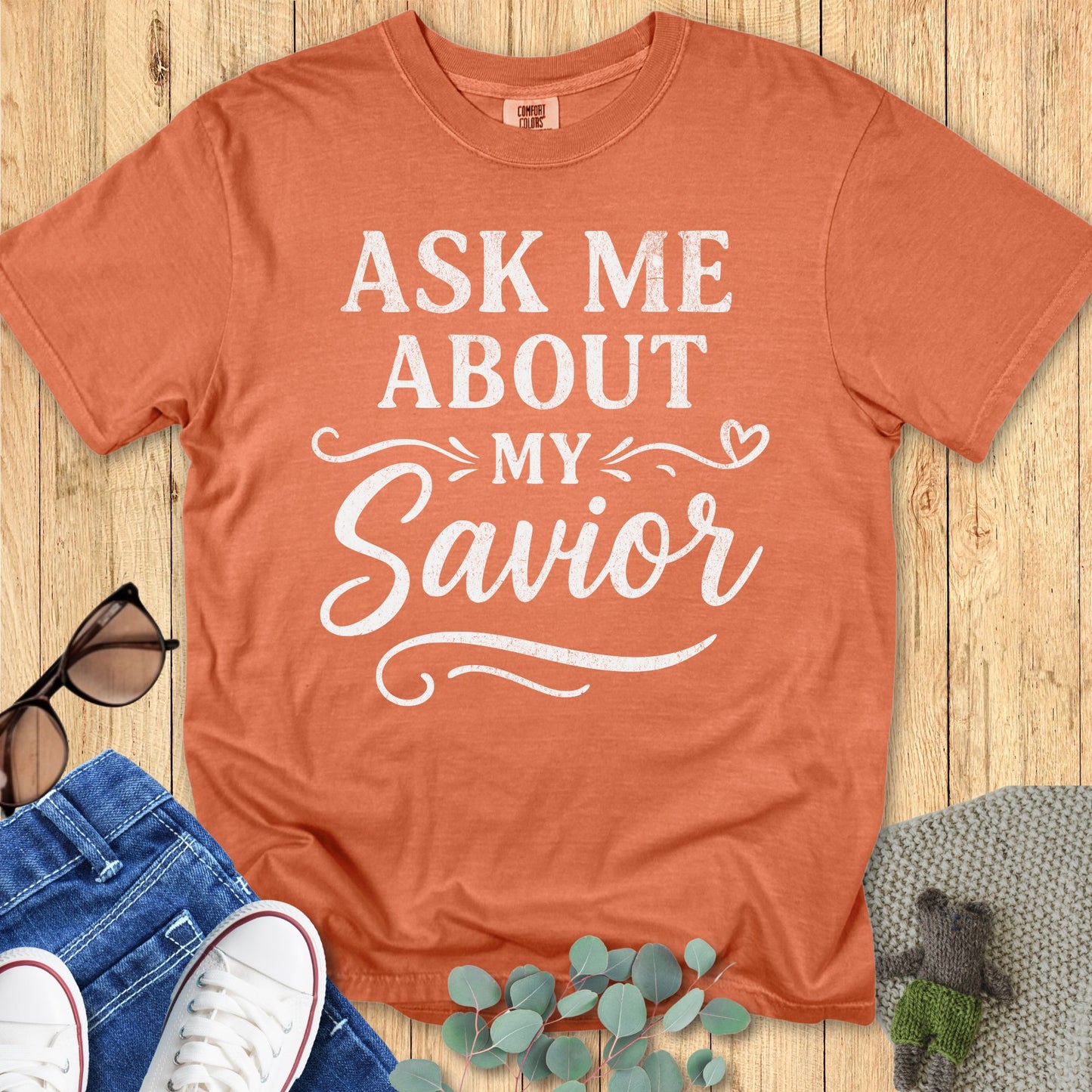 Terracotta Comfort Colors 1717 T-shirt with white vintage-style lettering that says “Ask Me About My Savior” in bold and elegant script, with decorative swirls and a heart motif. A soft Christian tee created to help women wear your faith daily. Shown flat on a wooden background with jeans, sneakers, sunglasses, and greenery.