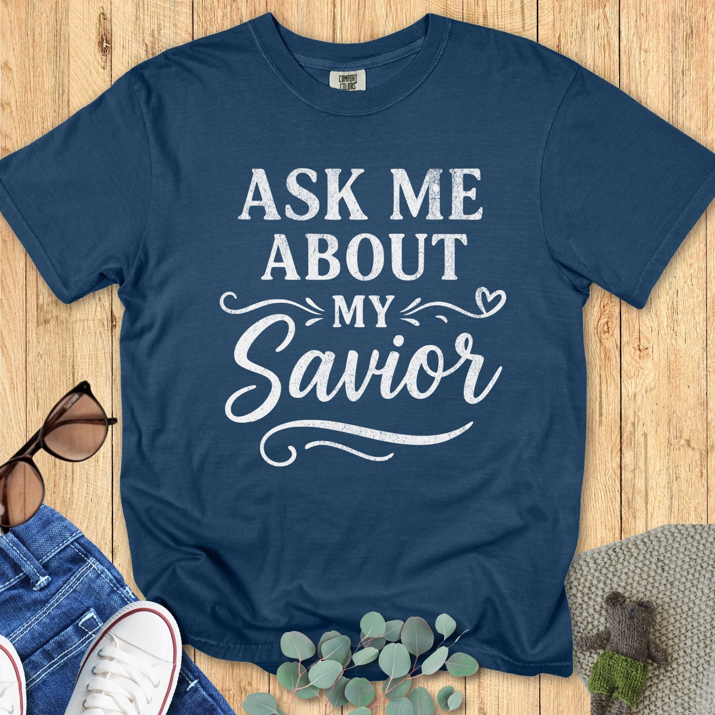 Midnight blue Comfort Colors 1717 T-shirt with white distressed lettering that reads “Ask Me About My Savior” in bold and script fonts, styled with decorative swirls and a heart accent. Christian graphic tee designed to help you wear your faith. Flat lay with jeans, sneakers, sunglasses, and greenery on a wooden background.