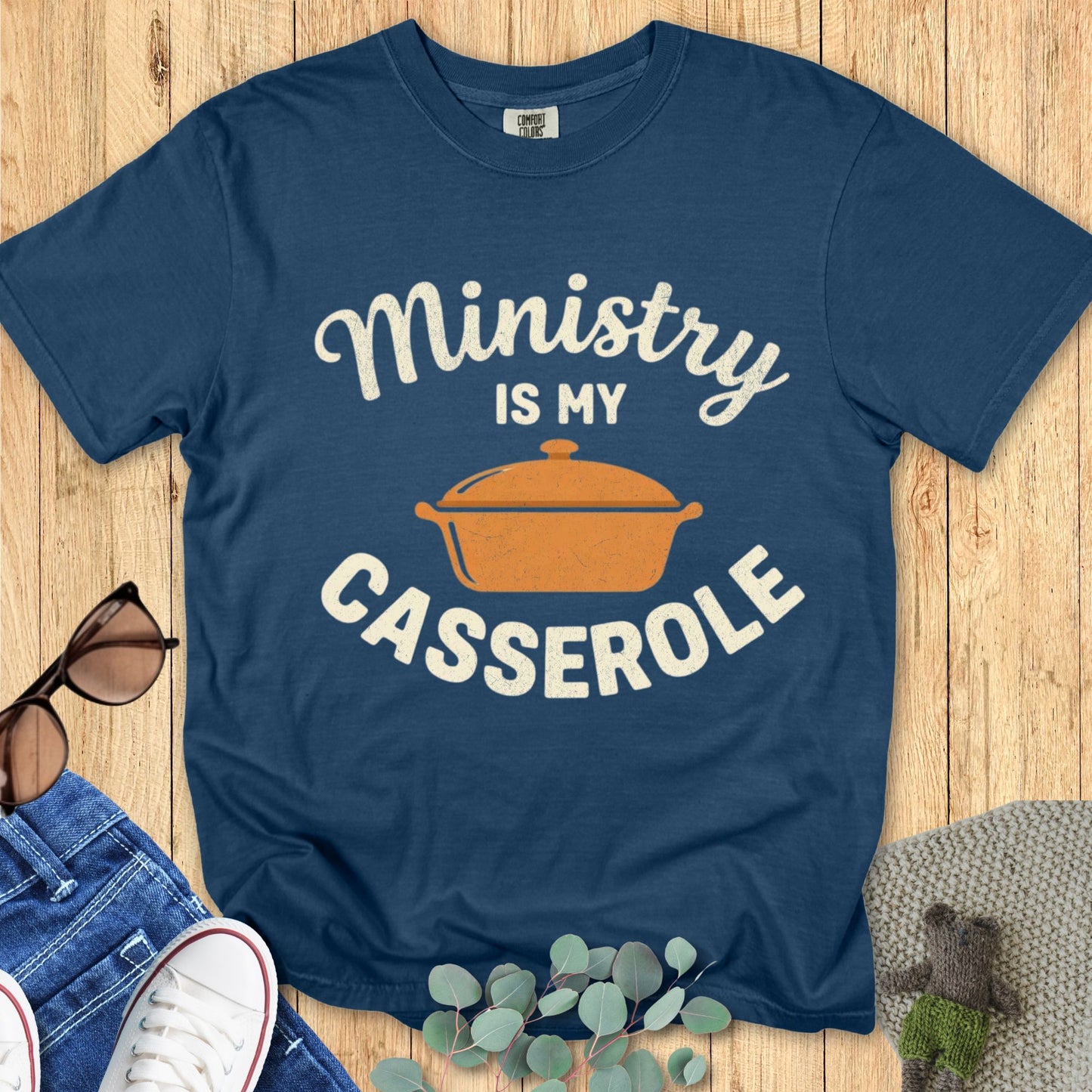 Blue t-shirt with 'Ministry is my Casserole' text and pot graphic on a wooden surface.