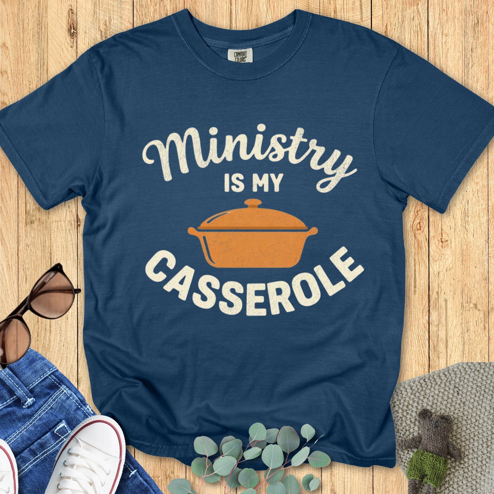 Blue t-shirt with 'Ministry is my Casserole' text and pot graphic on a wooden surface.