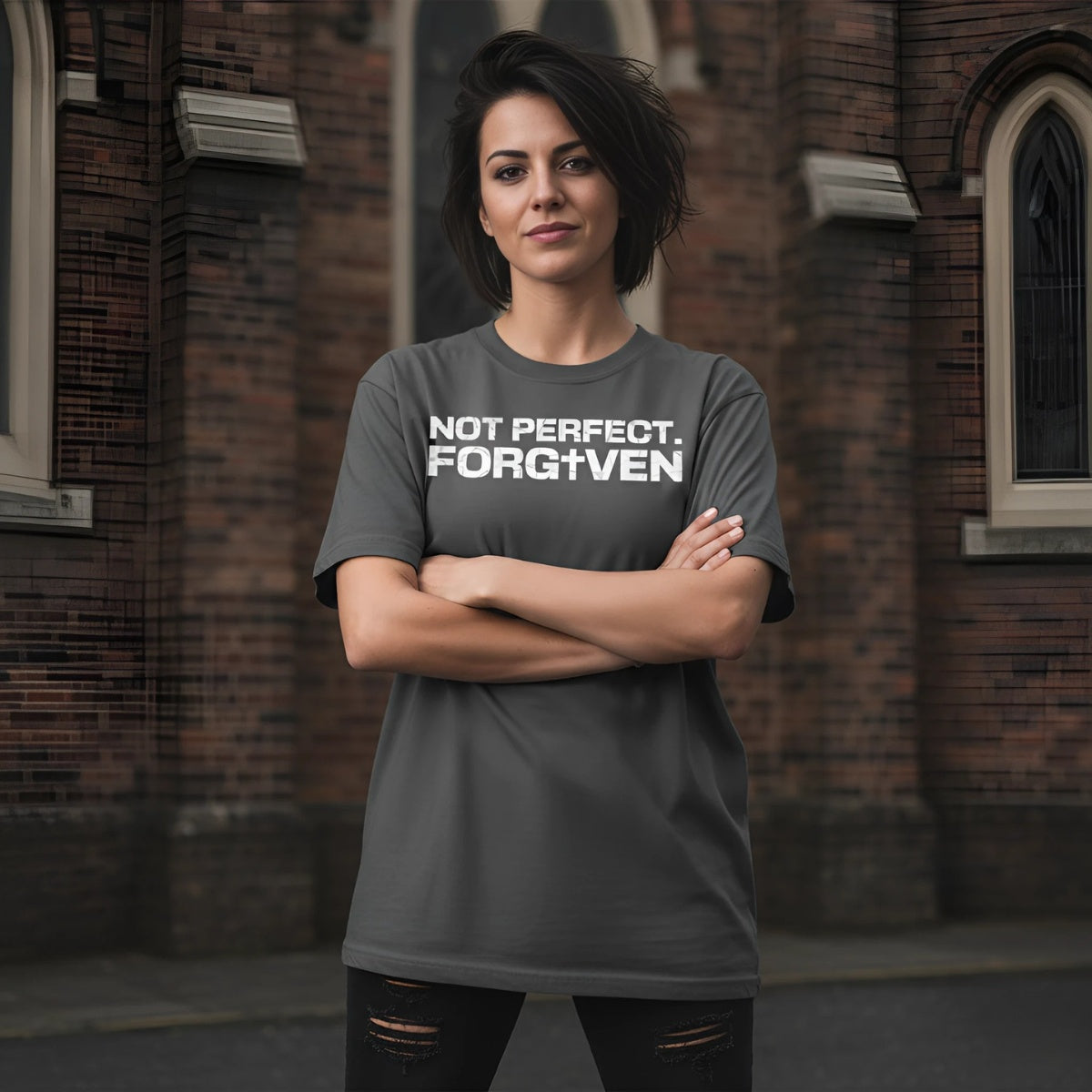 Person wearing a gray t-shirt with 'NOT PERFECT. FORGOTTEN' text in front of a brick building.