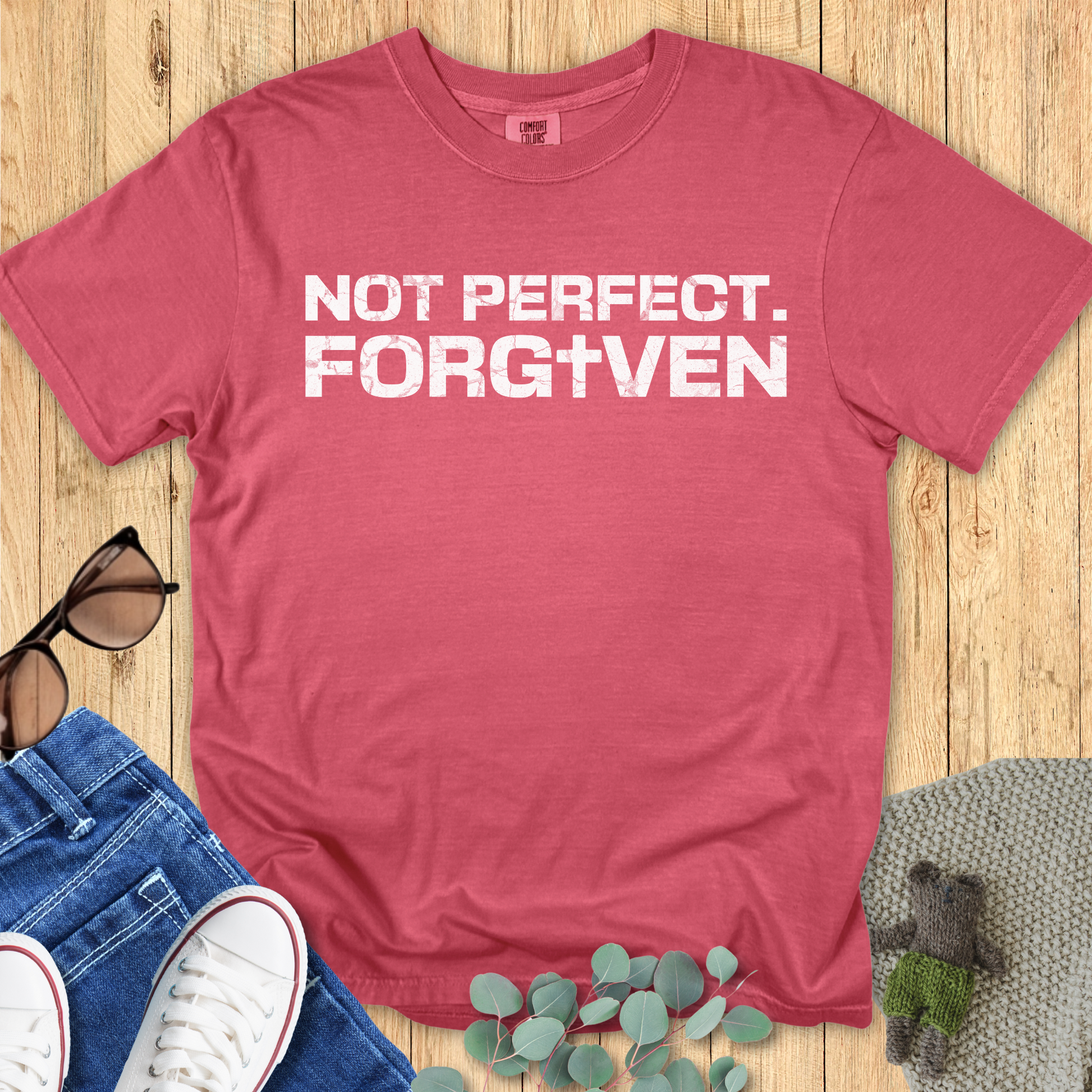 Red t-shirt with 'Not Perfect. Forgiven' text on a wooden surface with jeans, sneakers, and a plant.