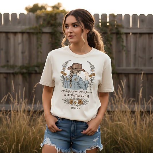 A woman wearing a Comfort Colors 1717 Christian women t-shirt in a natural outdoor setting. The shirt design features a rustic illustration of a woman in a cowboy hat surrounded by wildflowers, with the Bible verse โPerhaps you were born for such a time as this โ Esther 4:14โ printed below. The soft vintage style and relaxed fit highlight the boutique faith-based design.