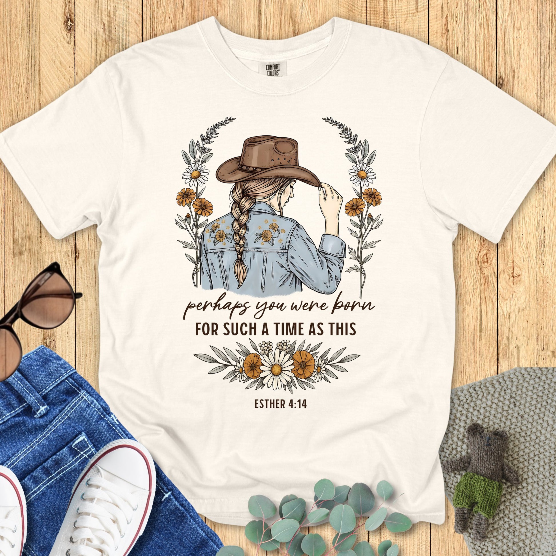 White t-shirt with a graphic of a woman wearing a cowboy hat and text on a wooden surface with accessories.