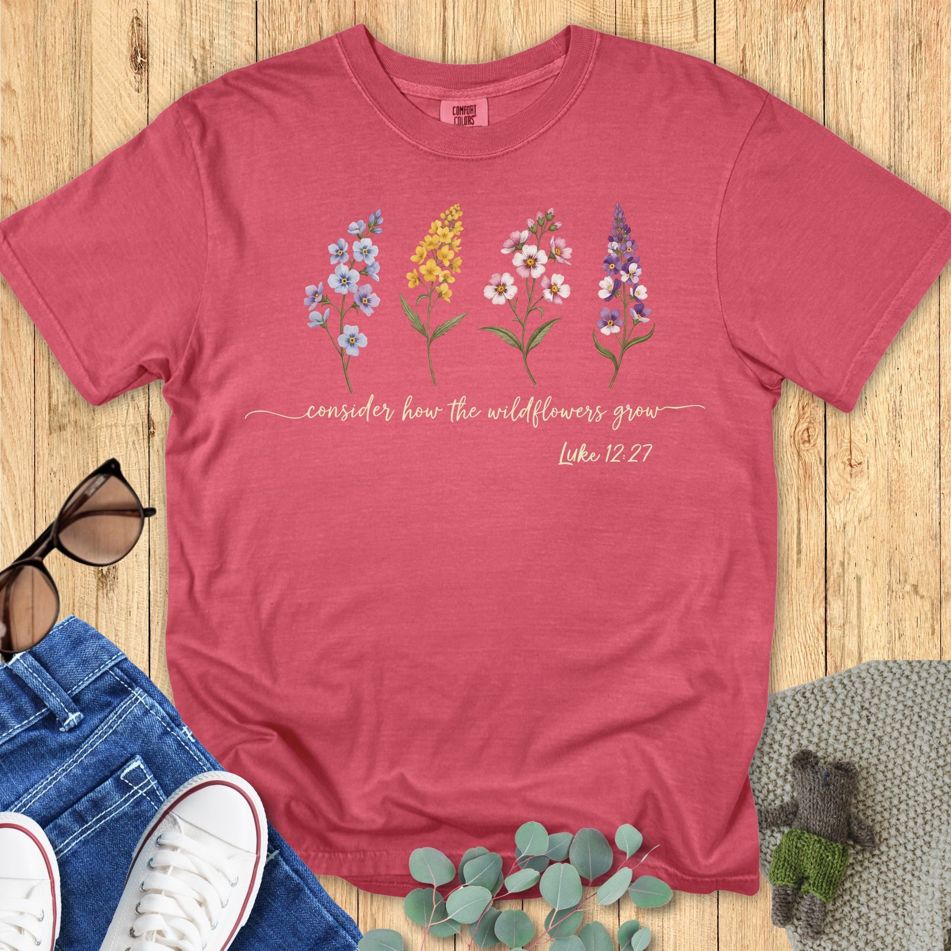 omfort Colors 1717 Christian women t-shirt in crimson flat lay. Christian floral design with wildflowers and Bible verse “Consider how the wildflowers grow – Luke 12:27” printed across the chest