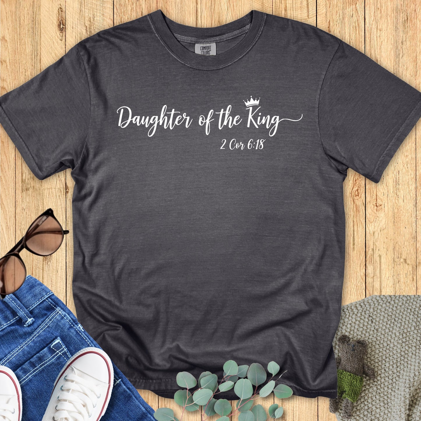Charcoal pepper Comfort Colors T-shirt with the phrase “Daughter of the King” in elegant white script with a small crown graphic, featuring 2 Corinthians 6:18 beneath. Flat lay style with jeans, white sneakers, sunglasses, and eucalyptus leaves on a wooden background