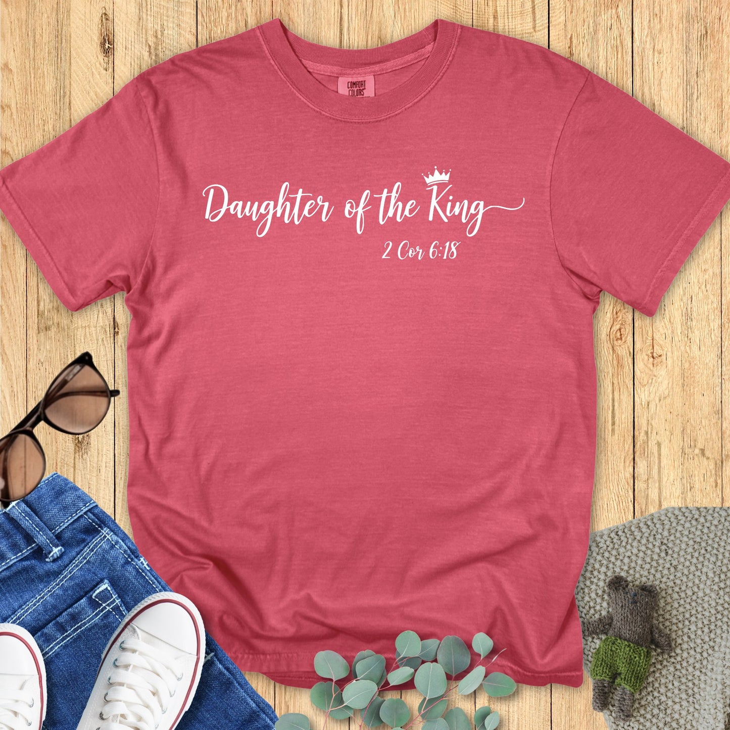 Crimson red Comfort Colors T-shirt with the phrase “Daughter of the King” in elegant white script with a small crown graphic, featuring 2 Corinthians 6:18 beneath. Flat lay style with jeans, white sneakers, sunglasses, and eucalyptus leaves on a wooden background