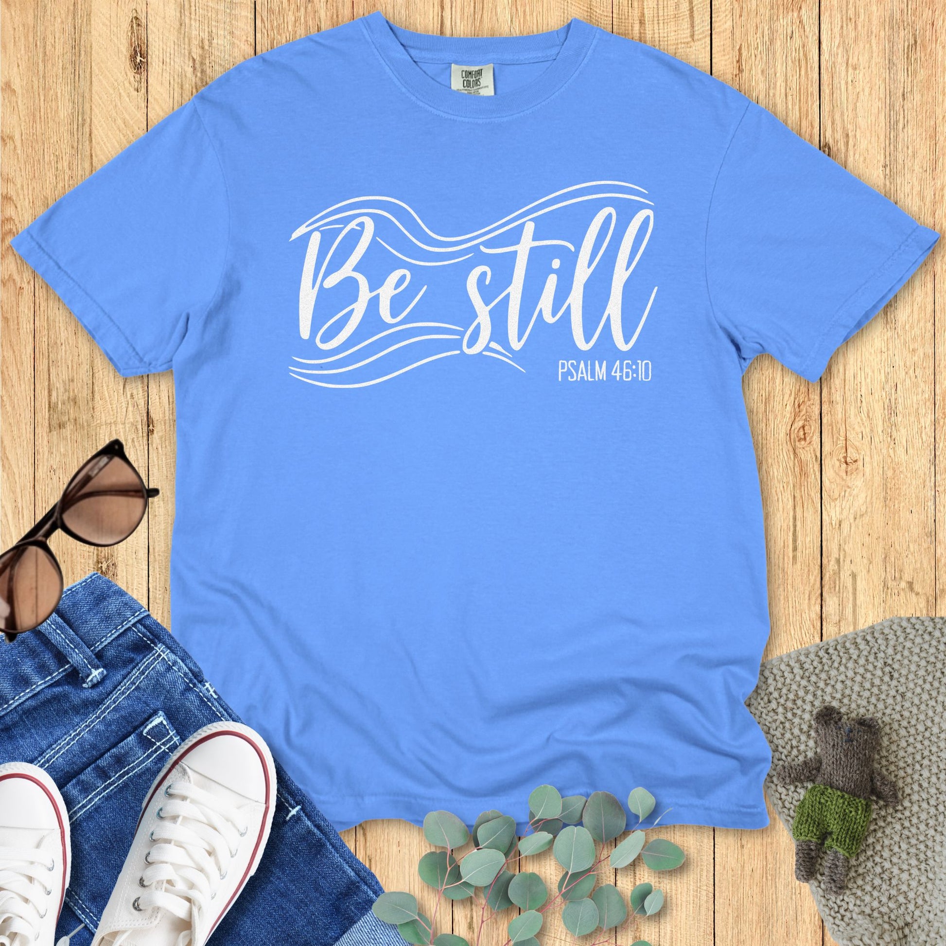 Christian T-shirt flat lay in mystic blue, featuring the words “Be Still” with flowing wave accents and “Psalm 46:10” in white script. Displayed on a wooden background with sunglasses, denim jeans, white sneakers, greenery, and a knitted blanket for styling