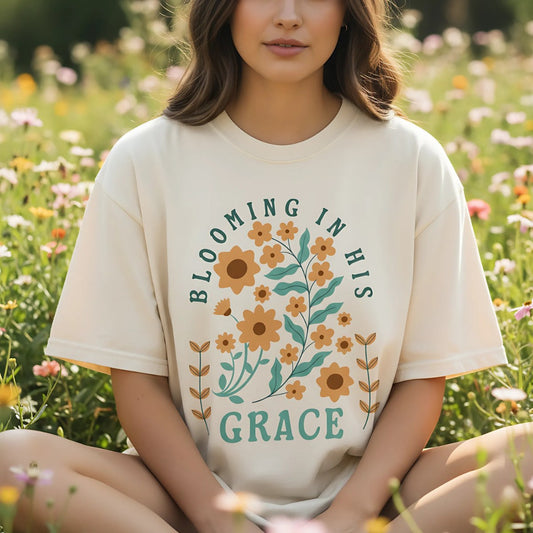 A Christian woman wearing a Comfort Colors 1717 Christian women t-shirt in cream with the design โBlooming in His Graceโ. The shirt features a faith-based floral print with orange flowers and teal lettering, photographed in a wildflower field