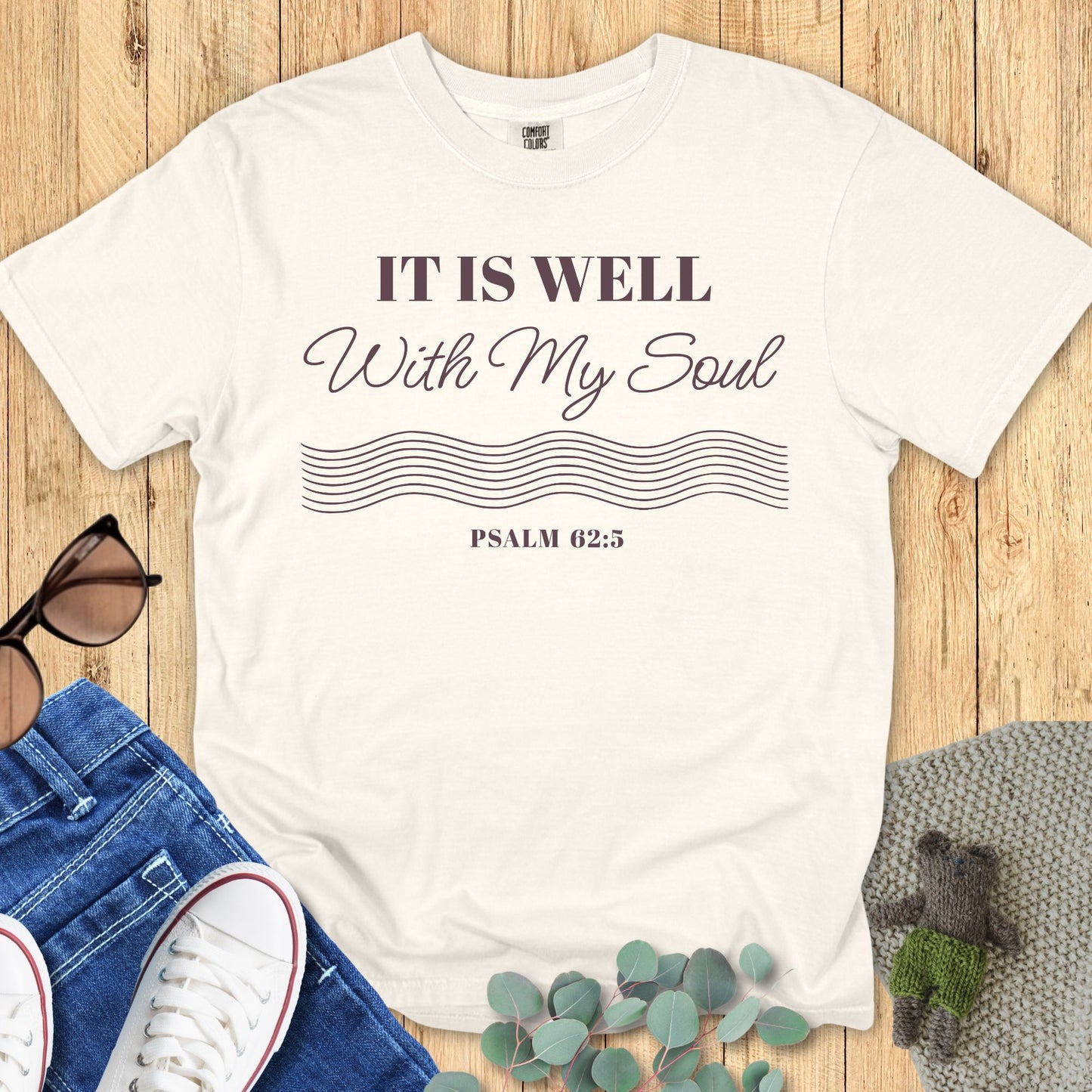Christian women’s t-shirt on Comfort Colors 1717 in ivory featuring the Bible verse ‘It Is Well With My Soul – Psalm 62:5’ with minimalist wave line graphic, styled in a flat lay with casual accessories