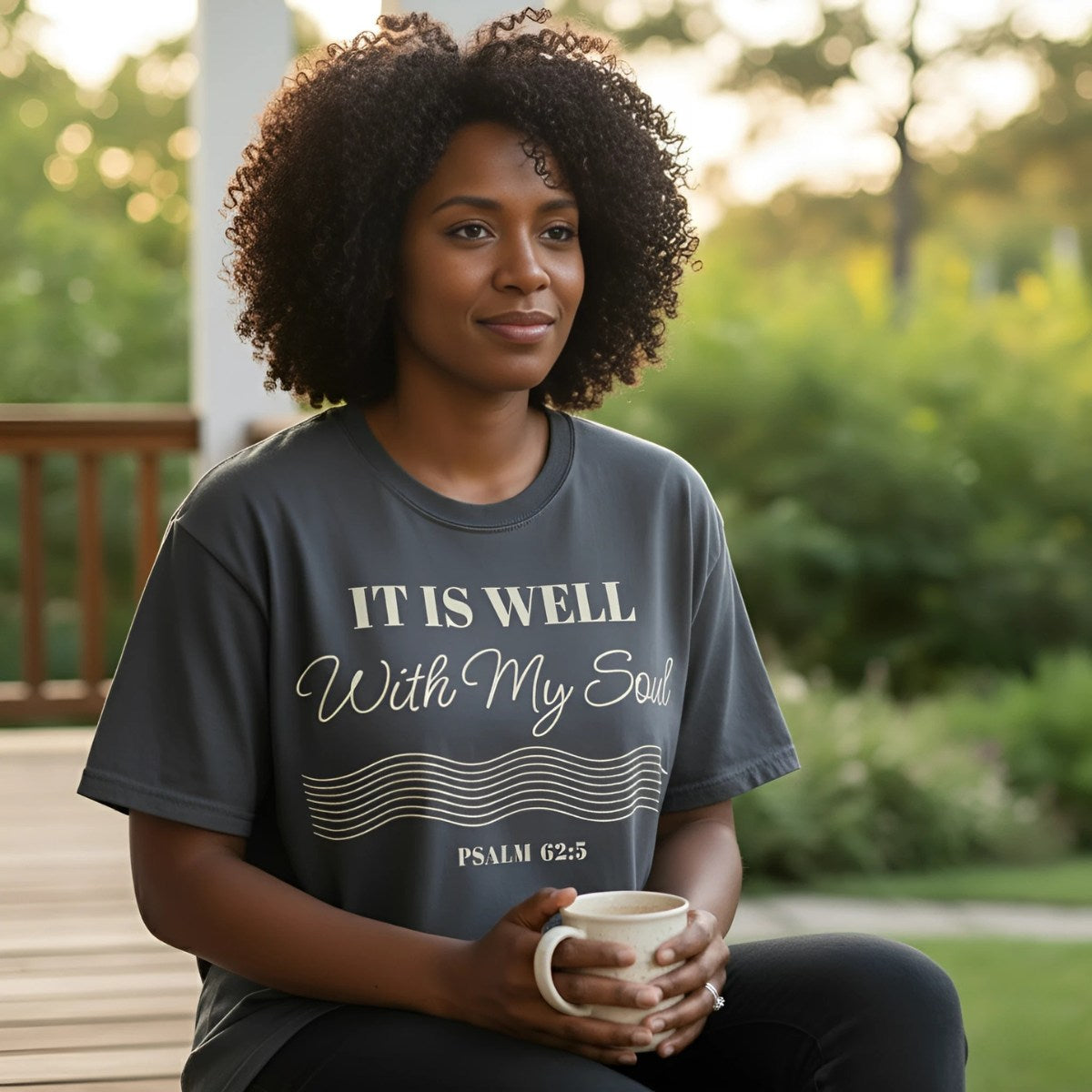 Smiling woman holding a mug while sitting outdoors in a pepper gray Comfort Colors 1717 Christian t-shirt that reads ‘It Is Well With My Soul – Psalm 62:5,’ a faith-based design with elegant script and wave lines