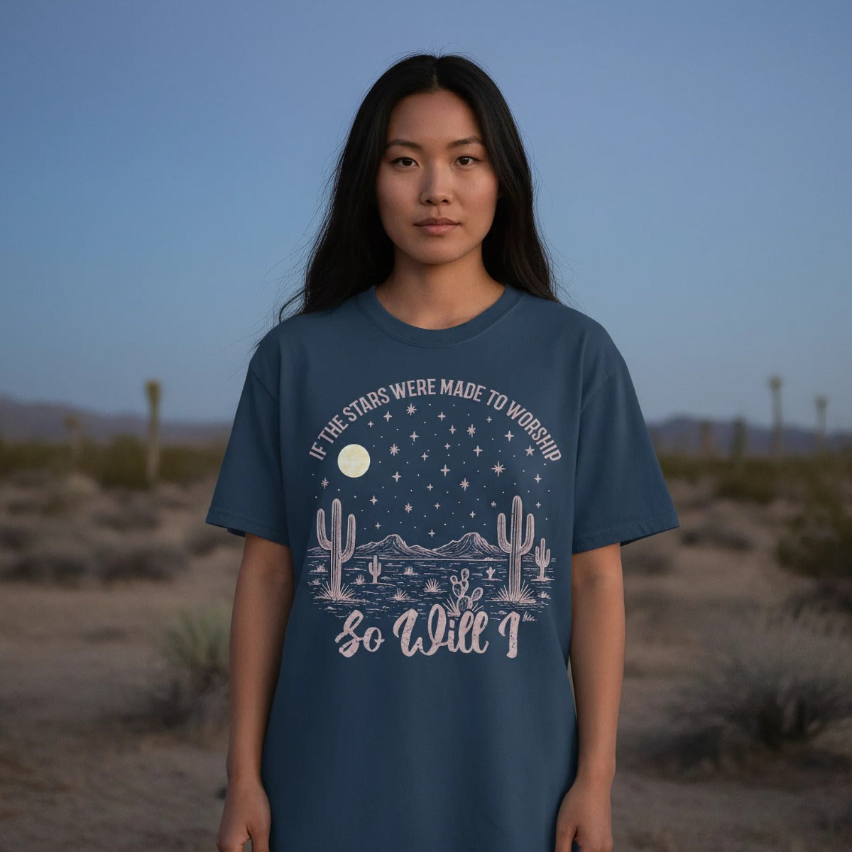 Young woman standing in the desert at dusk wearing a Christian women’s t-shirt that says 'If the stars were made to worship so will I,' featuring moon, stars, mountains, and cactus artwork