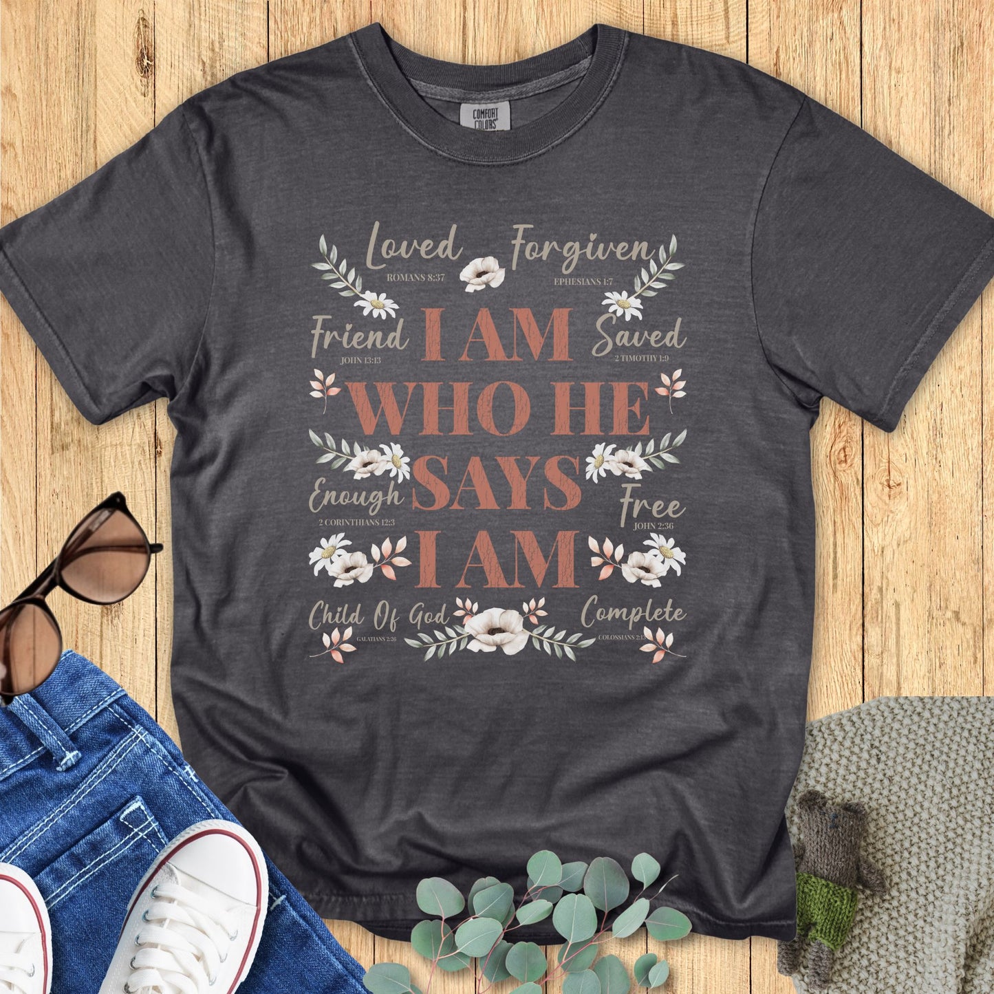 Christian women’s t-shirt in pepper gray with floral accents and the Bible-inspired affirmation “I Am Who He Says I Am” surrounded by words like Loved, Forgiven, Friend, Saved, Enough, Free, Child of God, and Complete. Styled with denim, sneakers, sunglasses, and eucalyptus on a wooden background