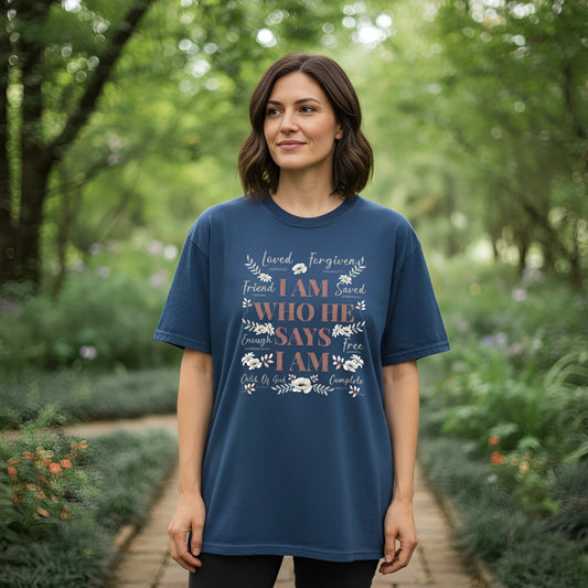 Woman standing on a garden path wearing a midnight blue Comfort Colors 1717 Christian t-shirt reading โI Am Who He Says I Am,โ decorated with soft white florals and scripture references, styled for everyday faith wear
