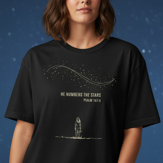 Woman wearing a black Comfort Colors 1717 Christian T-shirt against a starry night backdrop. The shirt design shows a girl gazing upward with the text โHe Numbers the Stars โ Psalm 147:4